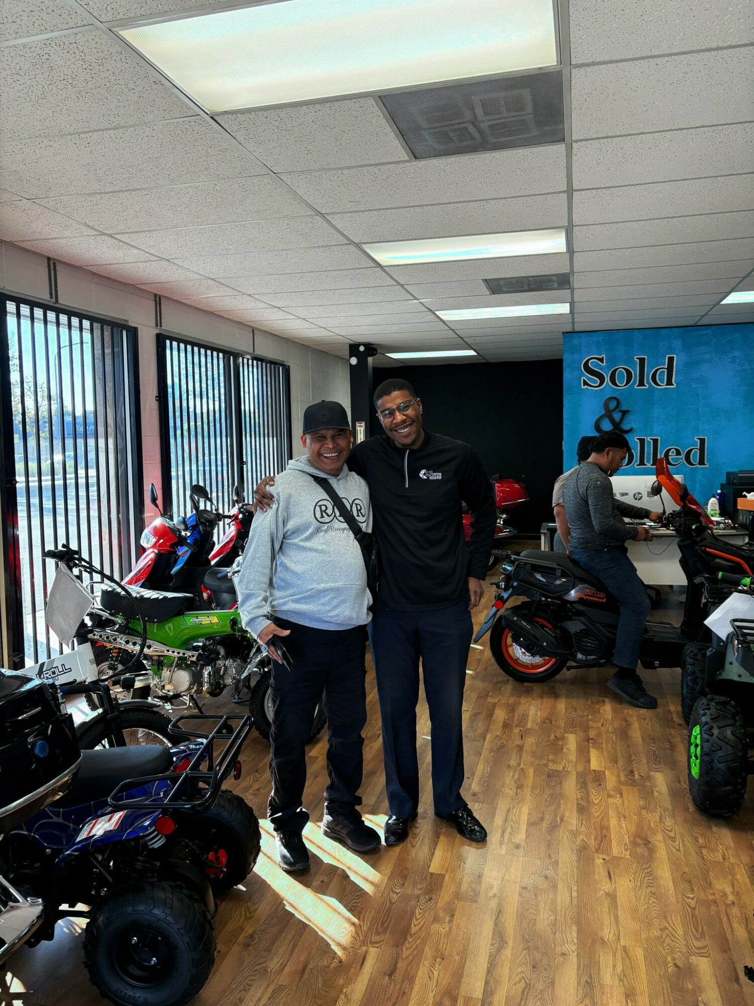 Two people standing inside a showroom with motorcycles, smiling, with large windows and a blue sign in the background.