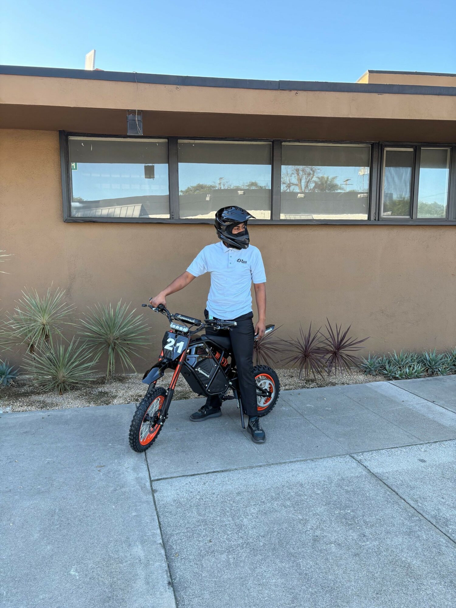 Person in white shirt and helmet standing next to a black and orange bicycle on sidewalk, building with large windows behind.
