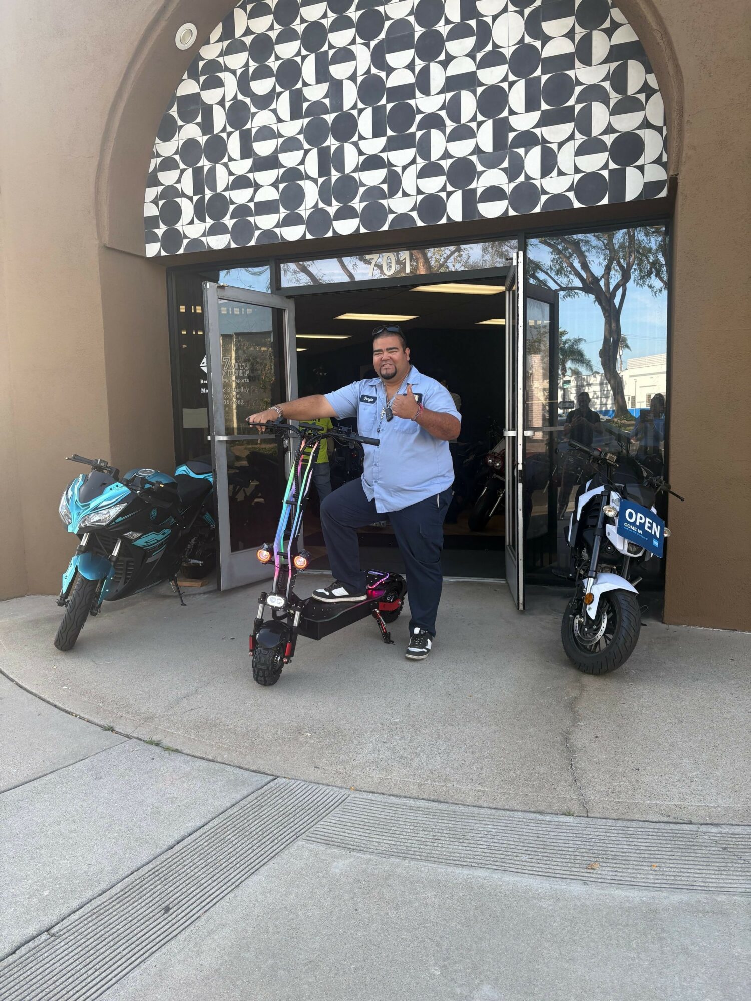Person standing outside a building with a scooter and a backpack on the sidewalk.