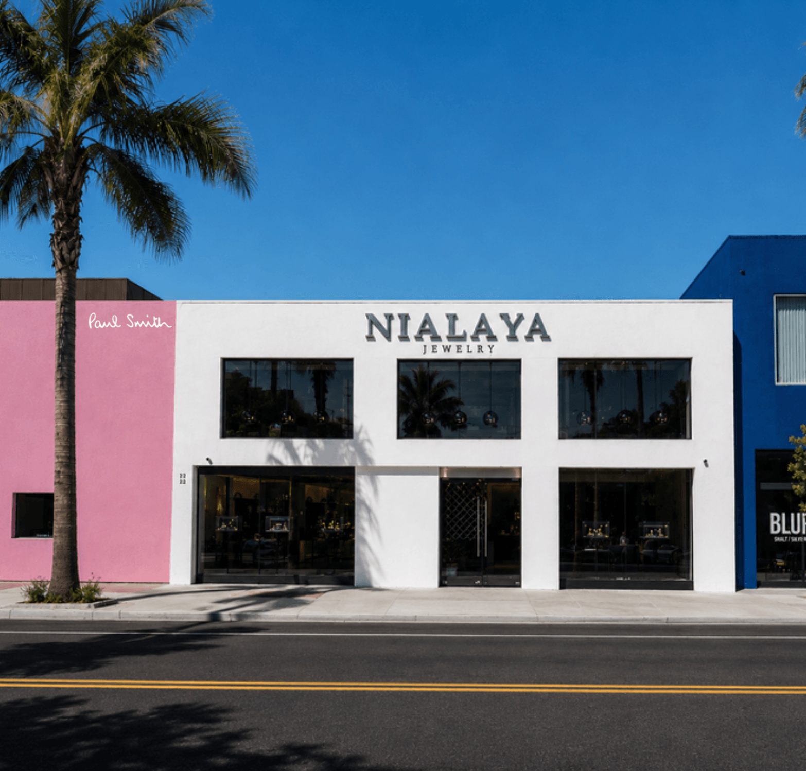 Storefront with large windows, sign reading 'NIALAYA JEWELRY', pink and white facade, palm trees, and a blue building nearby.