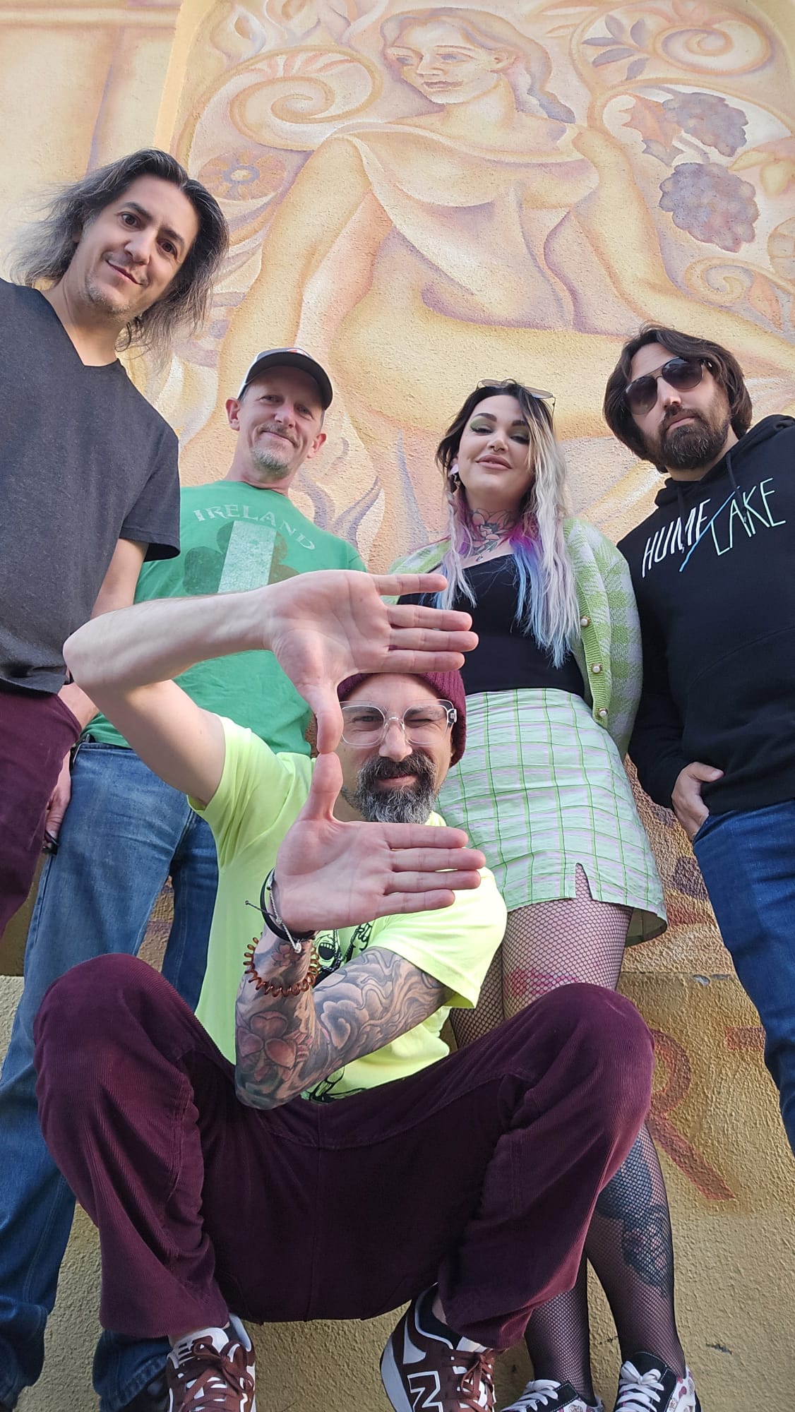 Group of five people standing outdoors in front of a large mural, one person sitting in front with tattoos and a beard.