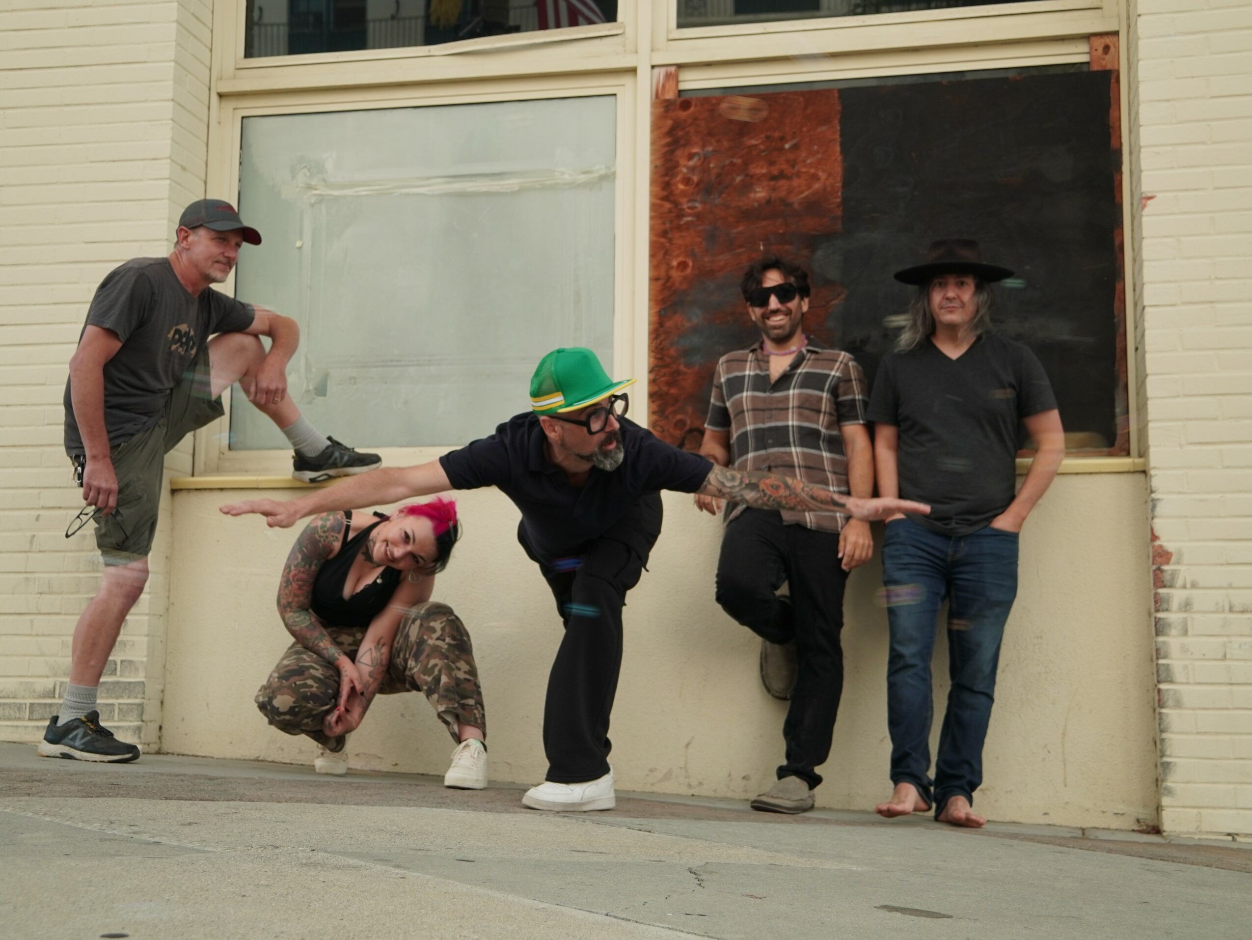 Five people standing and crouching in front of a building with a large window and a painted wall, some wearing hats and sunglasses.