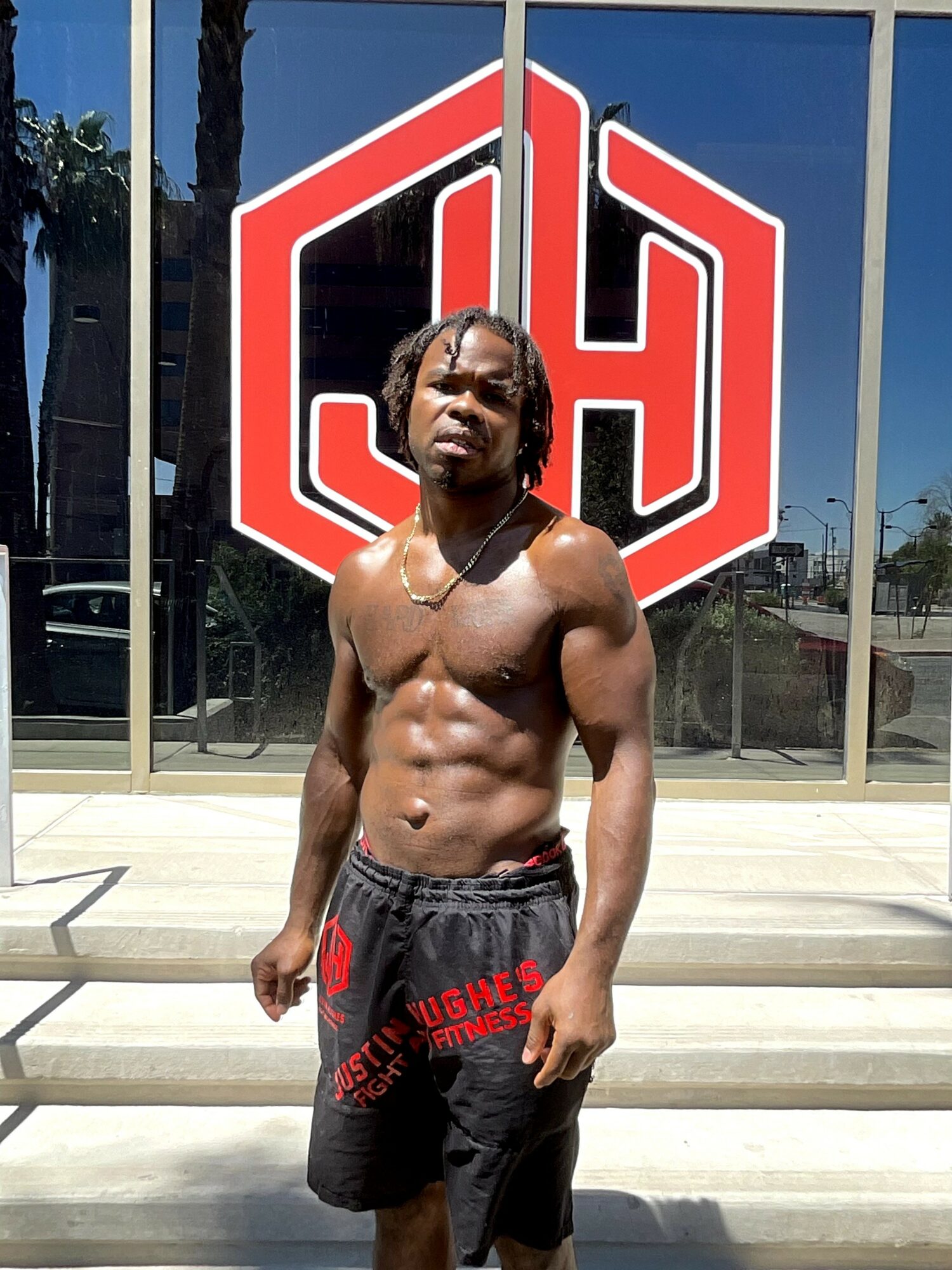 Young man with muscular build shirtless, standing outside in front of a glass building with a large red and white logo, sunlight casting shadows.