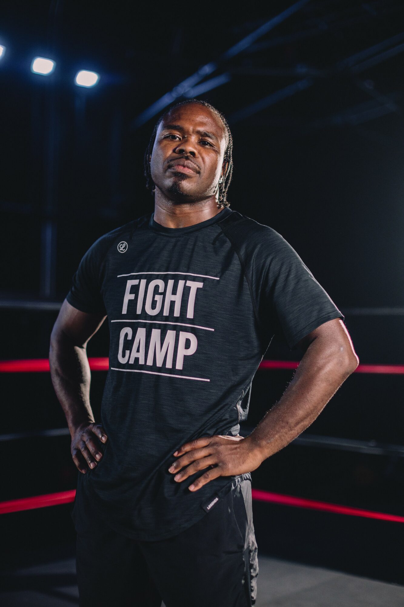 Man standing in a boxing ring with hands on hips, wearing a black T-shirt with 'FIGHT CAMP' text, in a dimly lit environment.