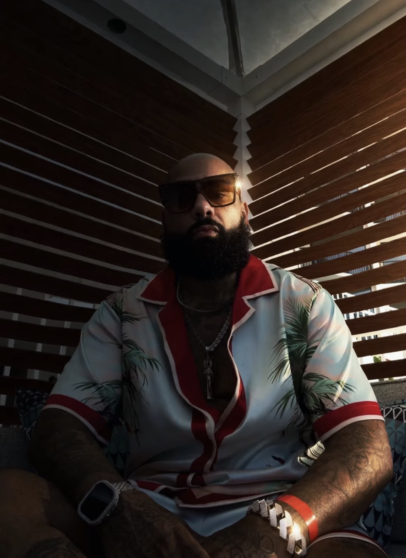 Man with beard wearing sunglasses, tropical shirt, and watch, sitting indoors with wooden blinds behind him.