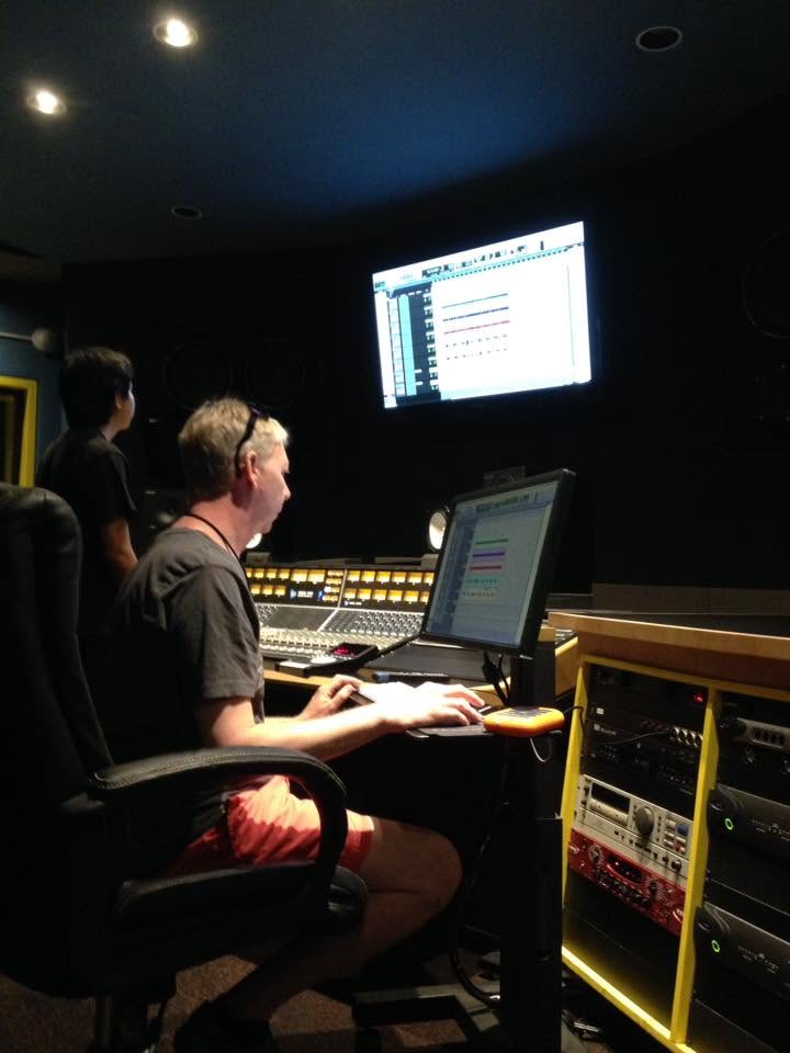 Person working at a mixing console with a computer in a dark recording studio, two people observing nearby.