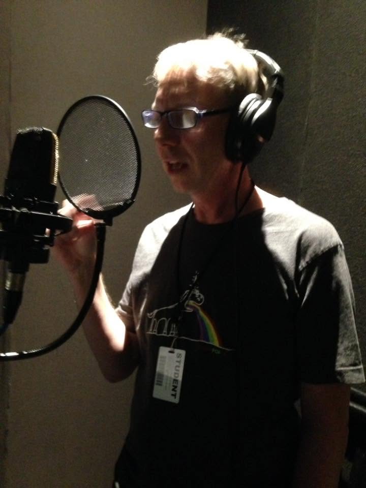 Person with glasses and headphones speaking into a microphone in a recording studio.
