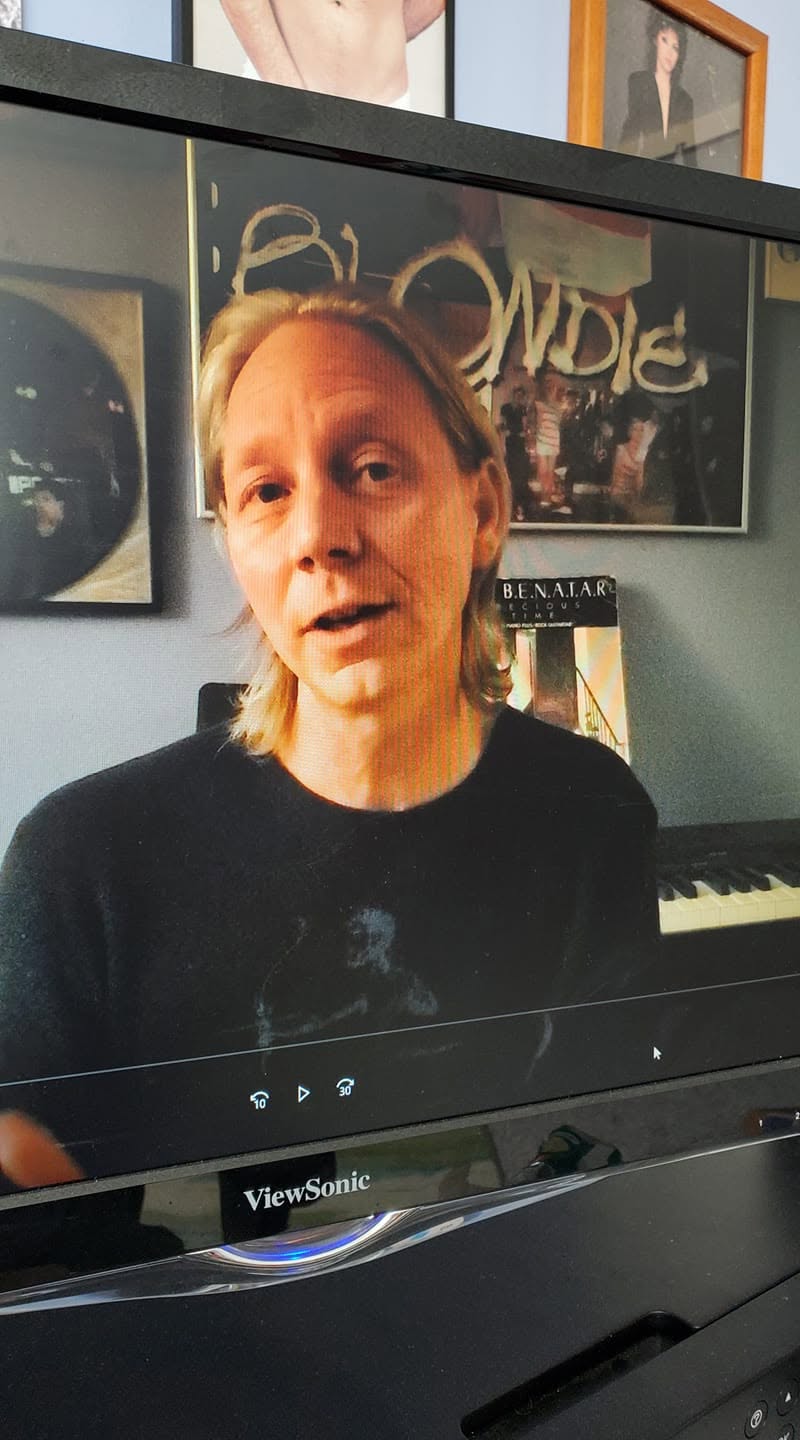 Person with shoulder-length hair and black shirt speaking, background includes framed pictures and a keyboard, indoors.