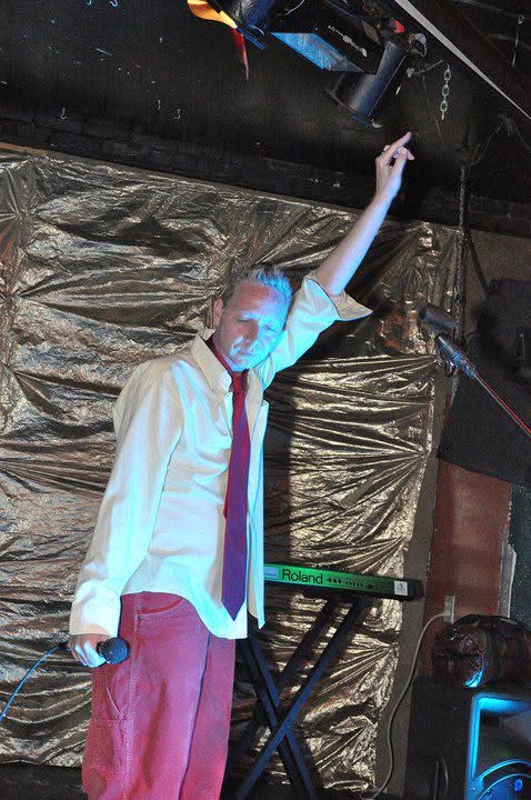 Person on stage with arm raised, wearing a white shirt and red pants, in front of a gold backdrop.