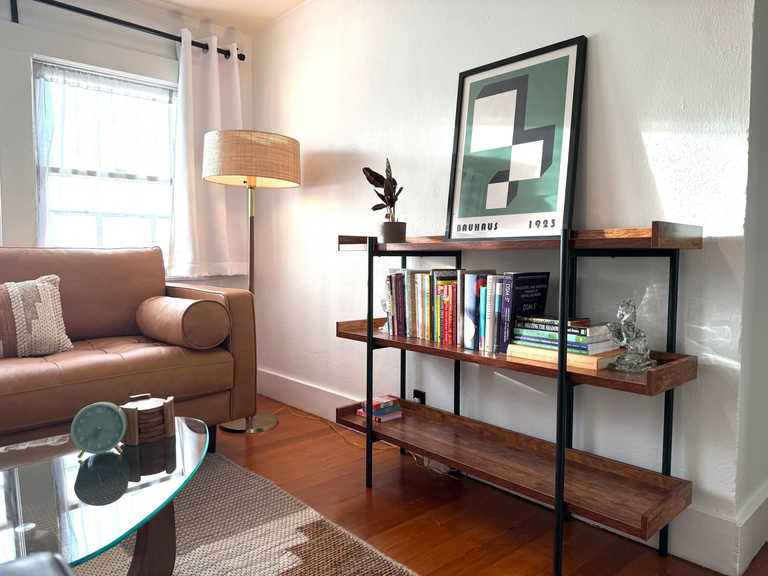 Living room with a beige sofa, a floor lamp, a bookshelf, and a framed artwork on a stand, with a window and curtains.