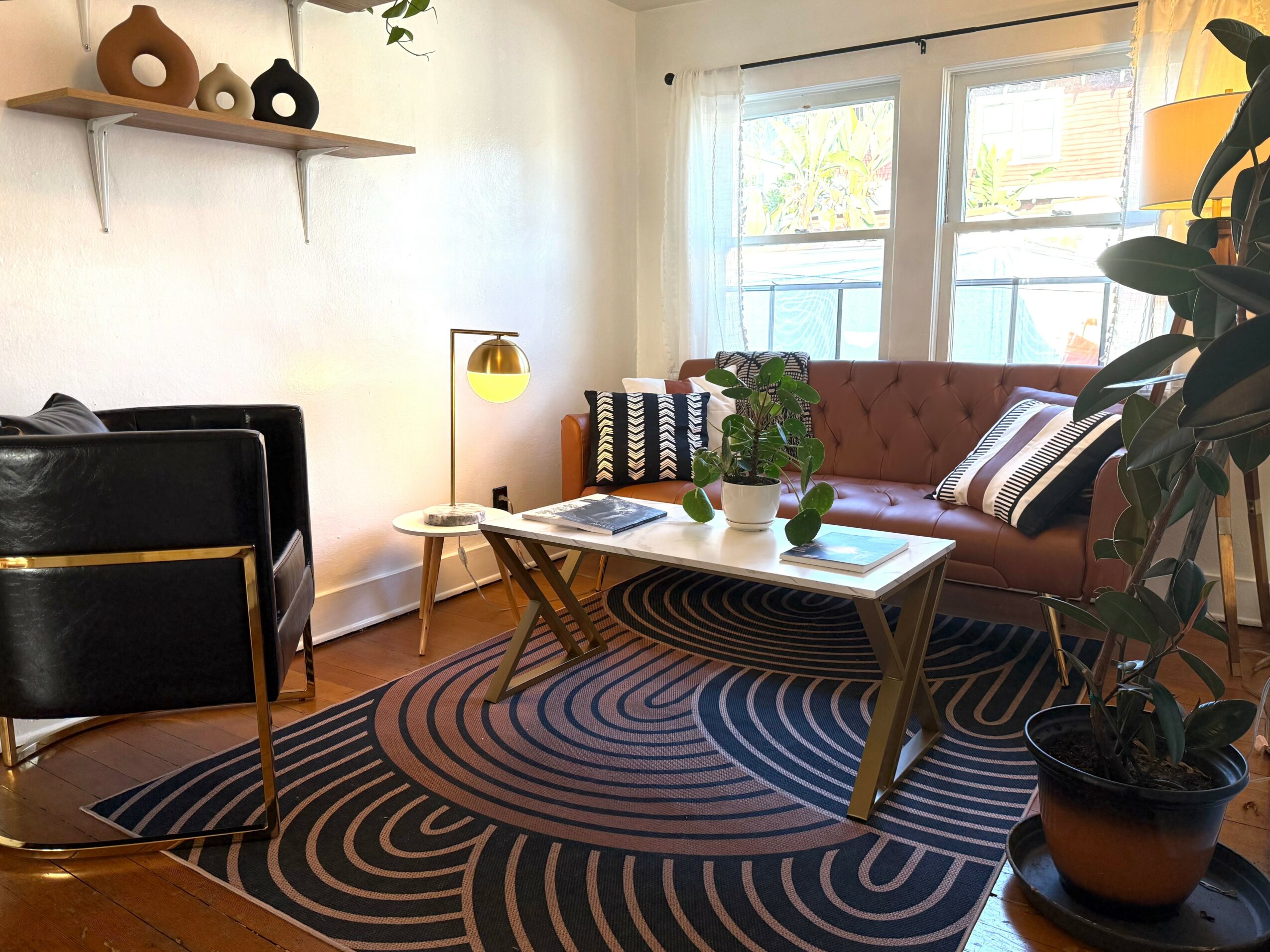 Living room with a pink sofa, black armchair, wooden coffee table, and a patterned rug. Large window with curtains, potted plant, and wall shelf.