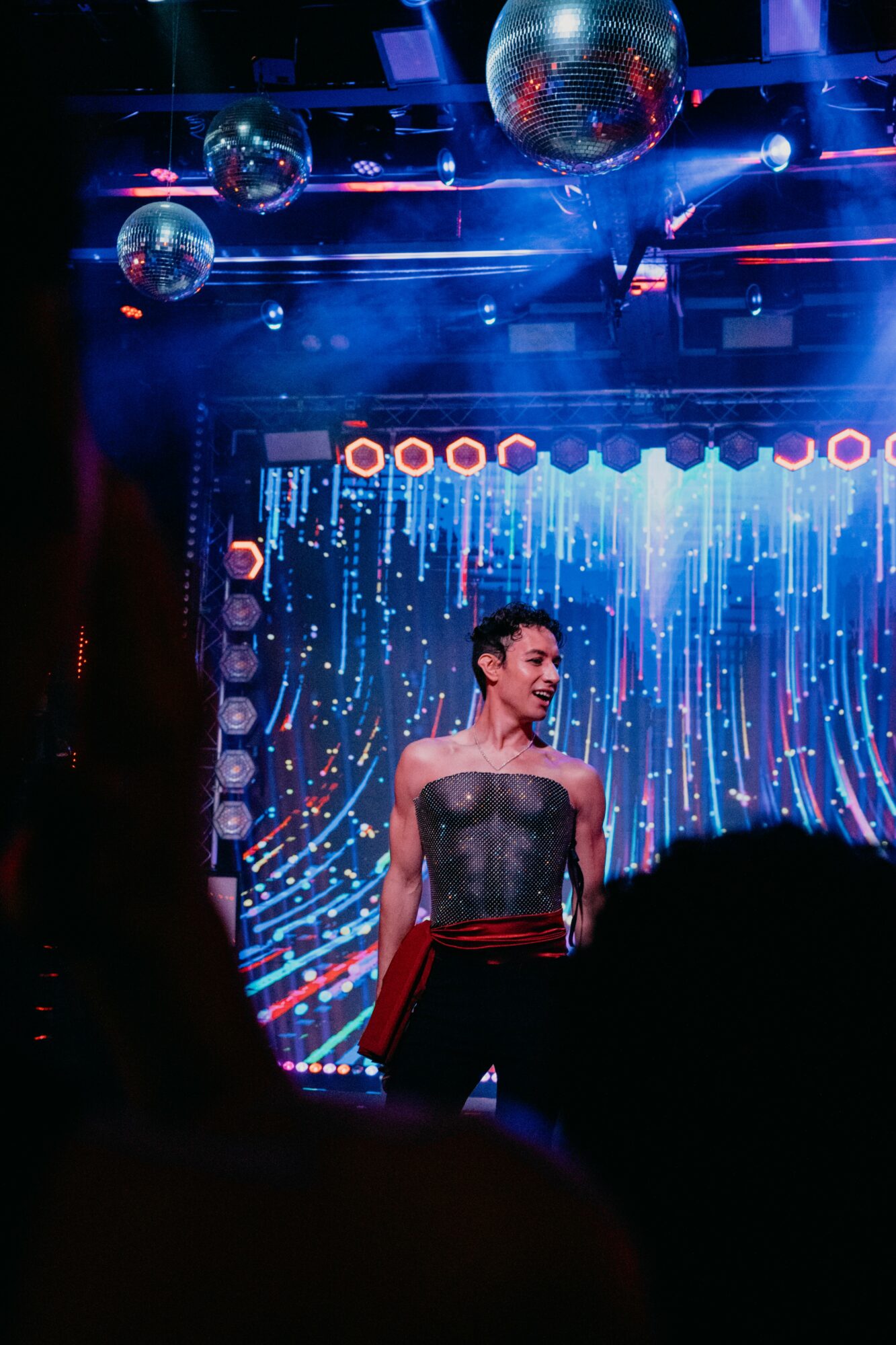 Performer on stage with a digital backdrop, surrounded by hanging disco balls, in a vibrant, colorful setting.