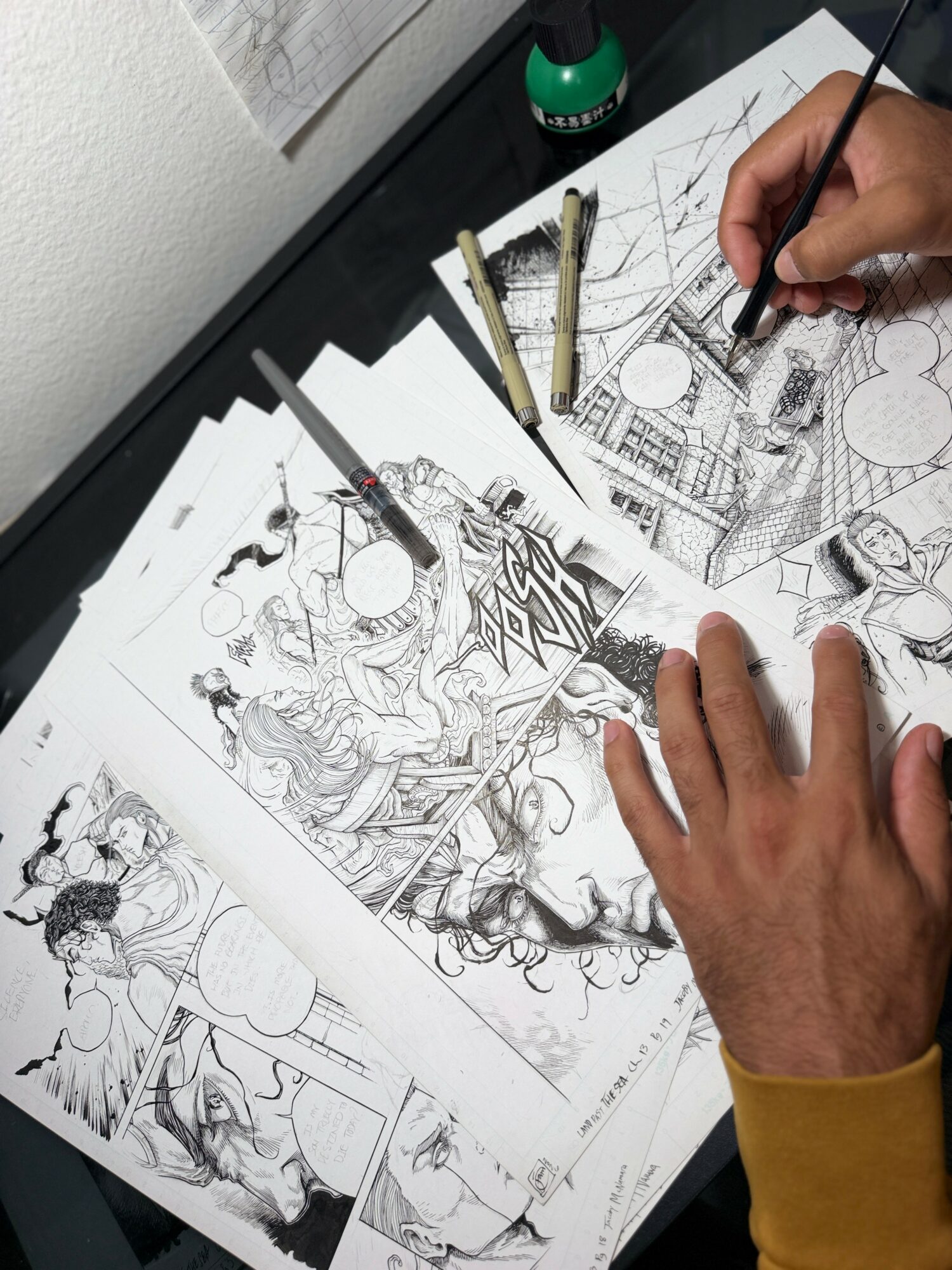 Person drawing detailed comic pages with pens and pencils on paper, workspace with ink bottle nearby.