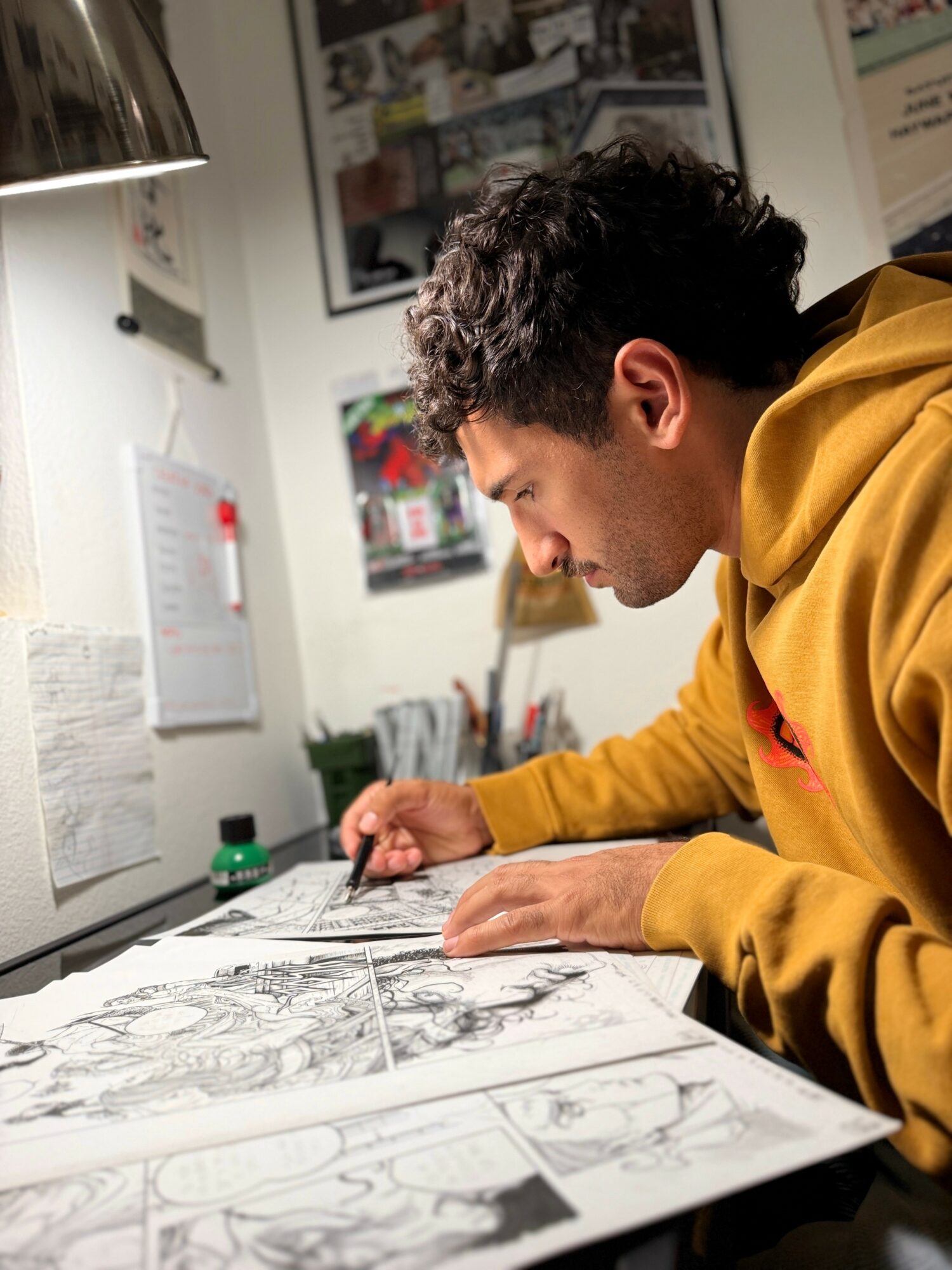 Young man with dark curly hair in a yellow hoodie working on comic book sketches at a desk, surrounded by papers and art supplies.