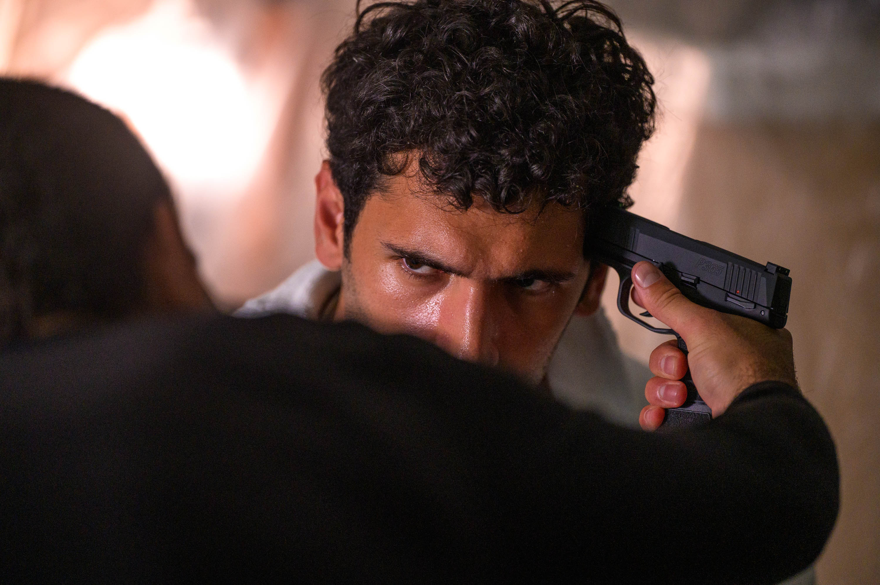 Person holding a gun to another person's head, with a focused expression, in a tense scene.