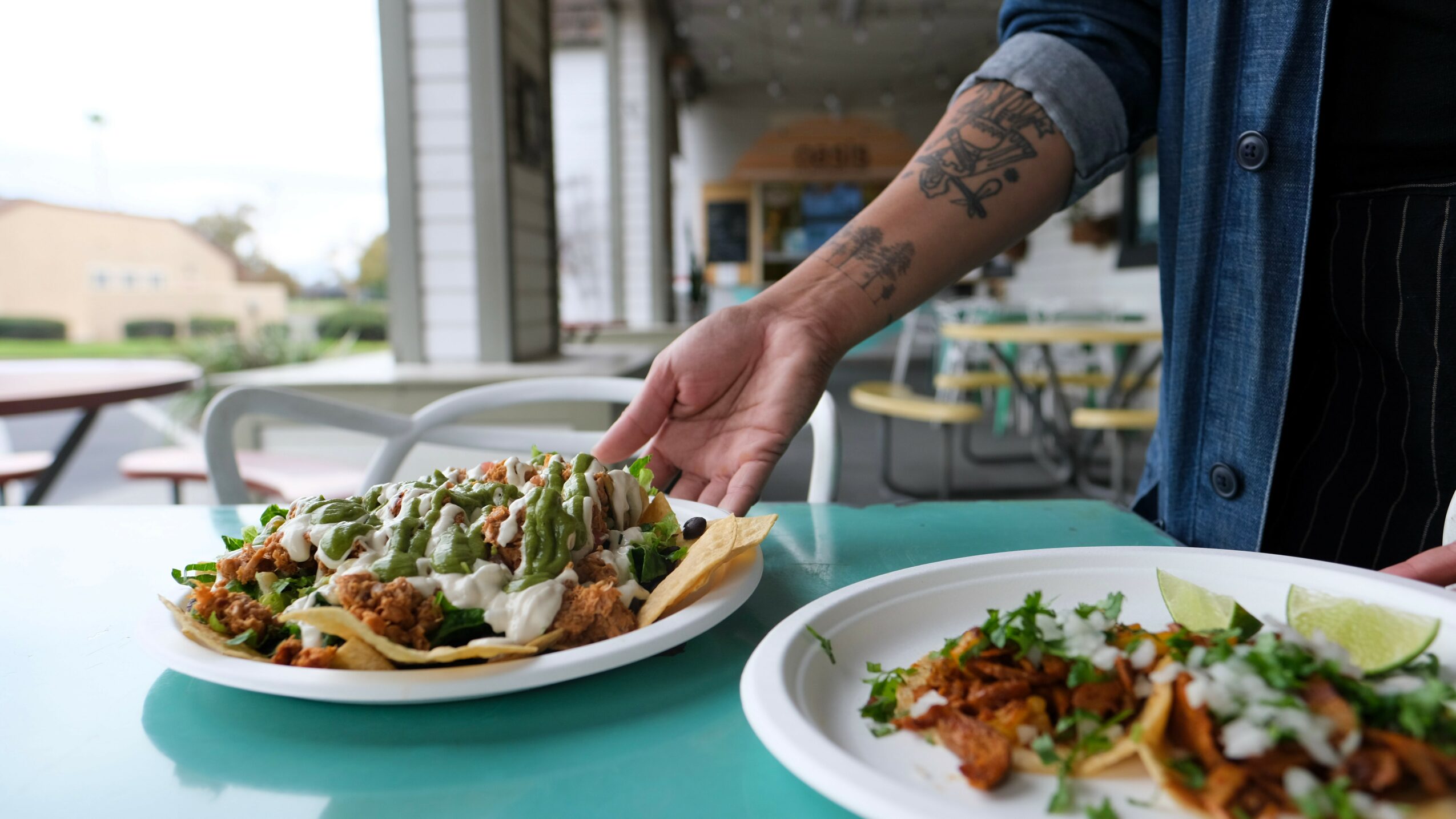 Person with tattooed arm reaching for food on a table outdoors, two plates of food visible, background of a patio area.