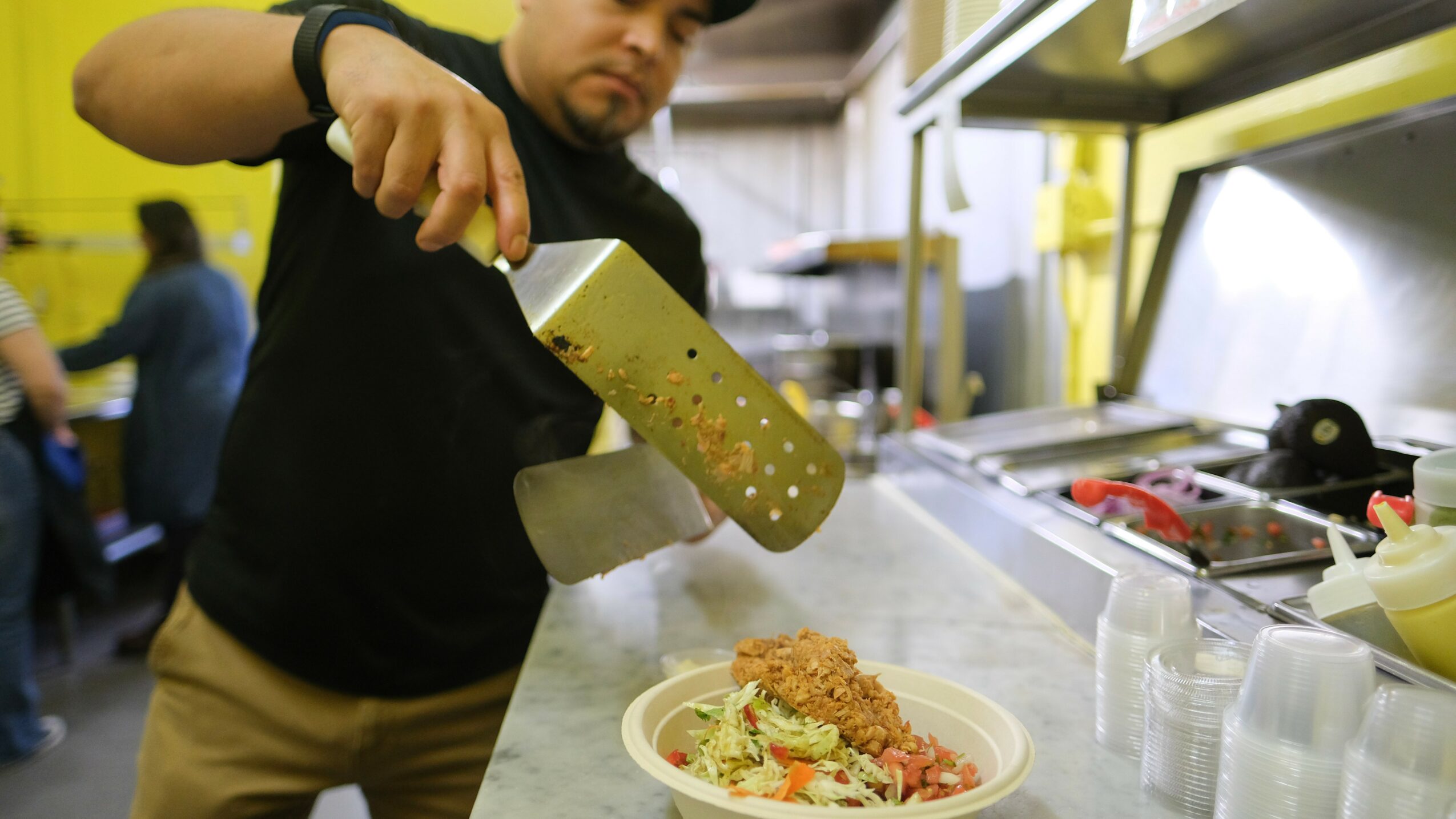 Person pouring sauce onto a plate of food in a kitchen, with a yellow wall in the background.