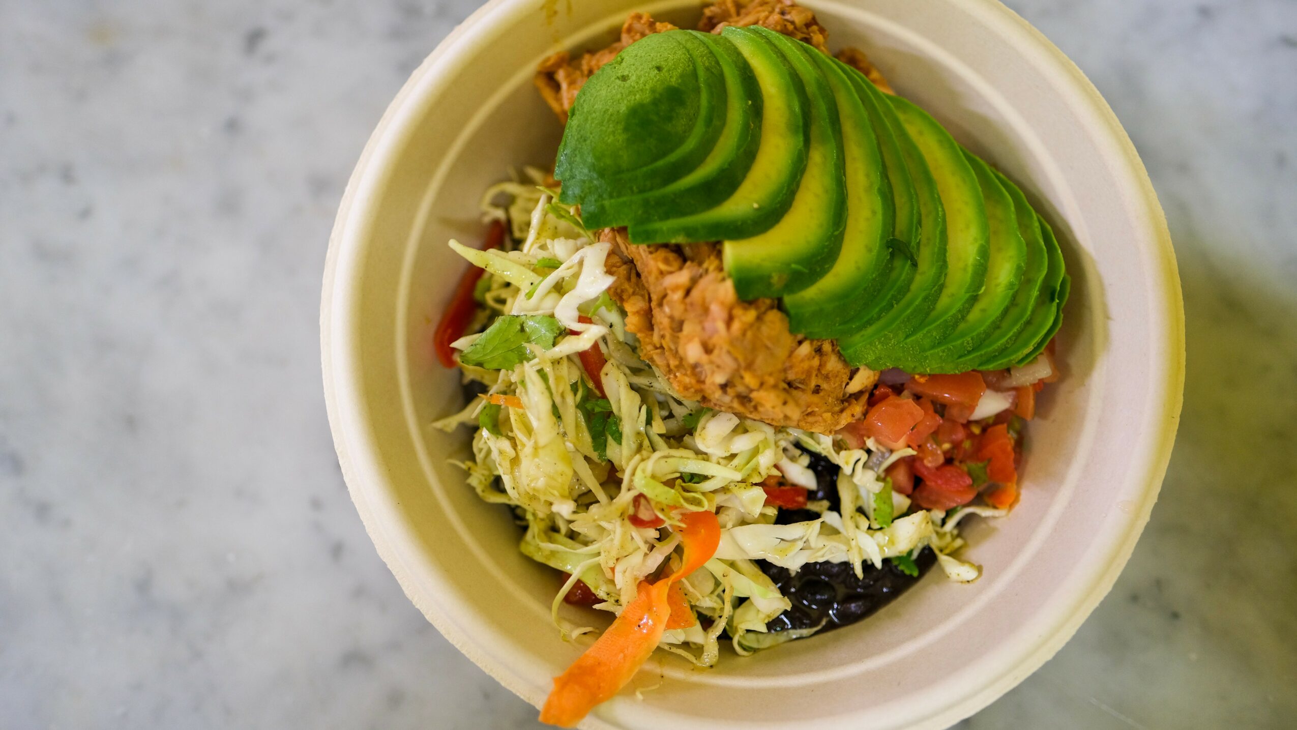 Bowl with sliced avocado, shredded lettuce, tomato, and other vegetables on a marble surface.