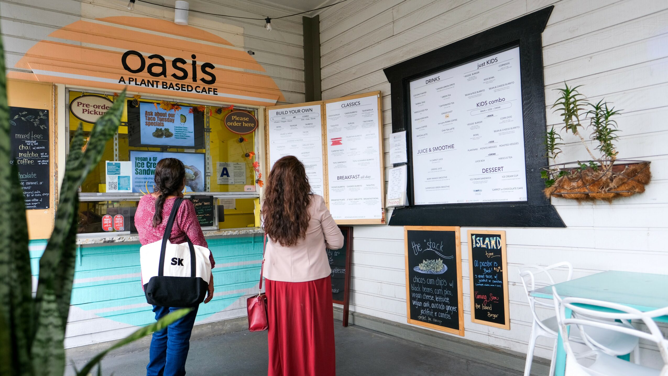 Two women look at a menu outside a cafe named Oasis, with a white wall and potted plant nearby.