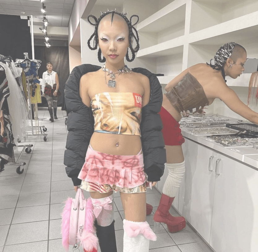 Young woman with unique hairstyle and fashion pose in store, wearing colorful crop top, pink skirt, and accessories.