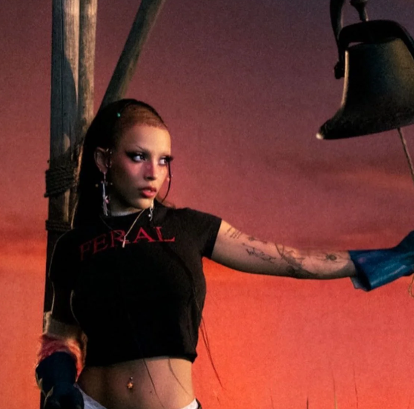 Young woman with long hair, wearing a black crop top, holding a bell, against a sunset sky.