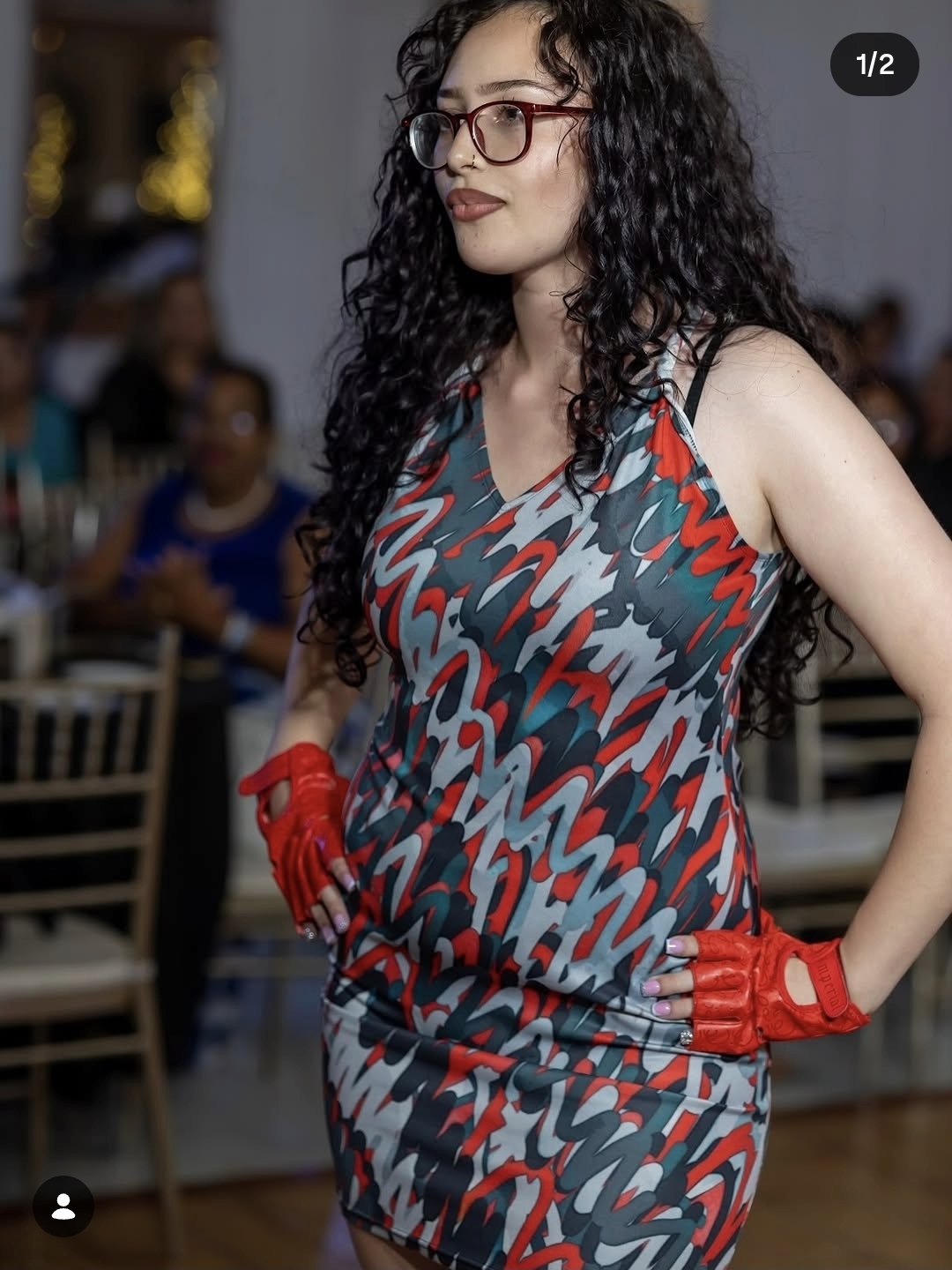 Woman with curly hair and glasses wearing a patterned dress and red gloves, standing indoors.