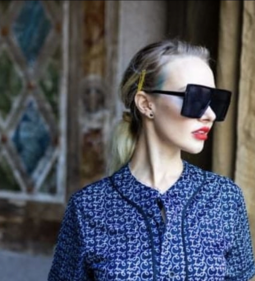 Woman with blonde hair, large sunglasses, red lipstick, wearing a blue patterned shirt, standing outdoors near wooden structures.