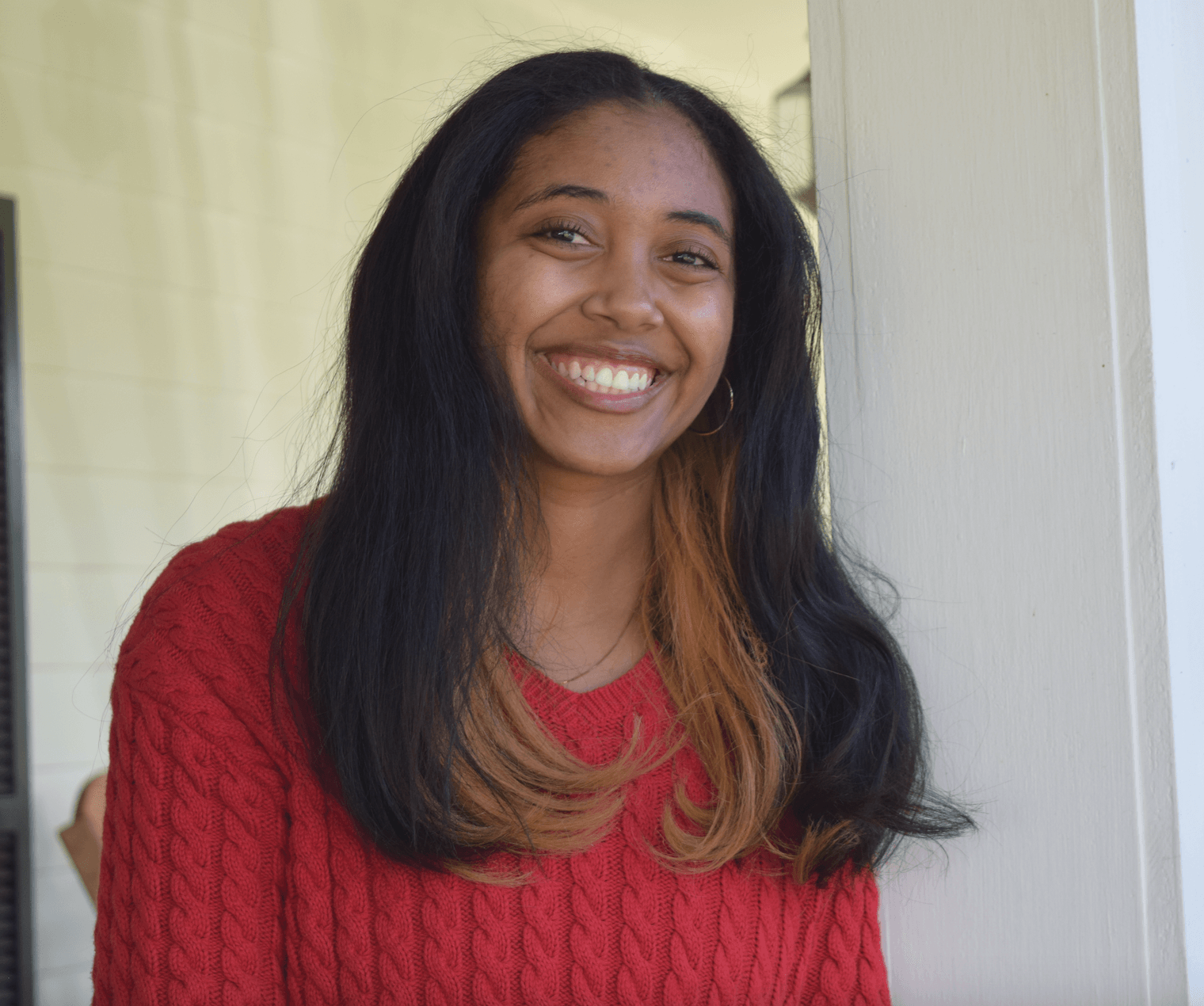 Smiling woman with long dark hair and blonde tips wearing a red sweater, standing indoors near a window.