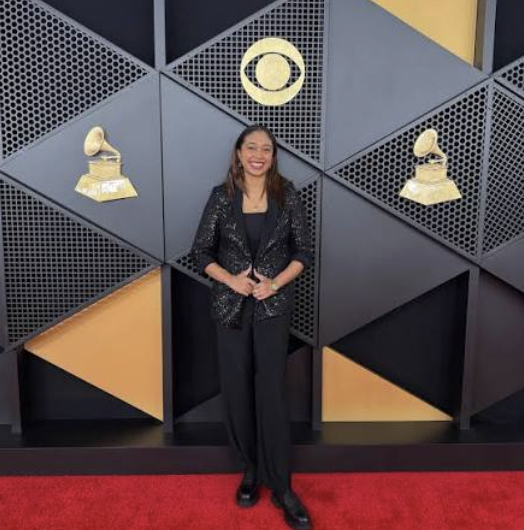 Woman standing on red carpet in front of geometric wall with Grammy awards and CBS logo, smiling, wearing black outfit.