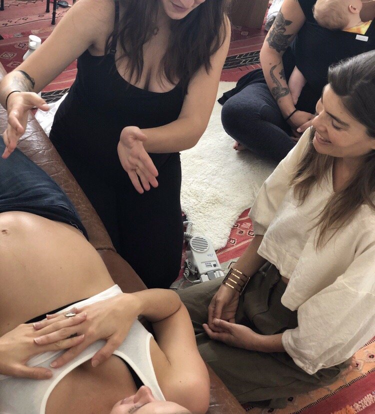 Group of women gathered around a person lying on a table, with one woman touching the person's abdomen, in a room with a patterned carpet.