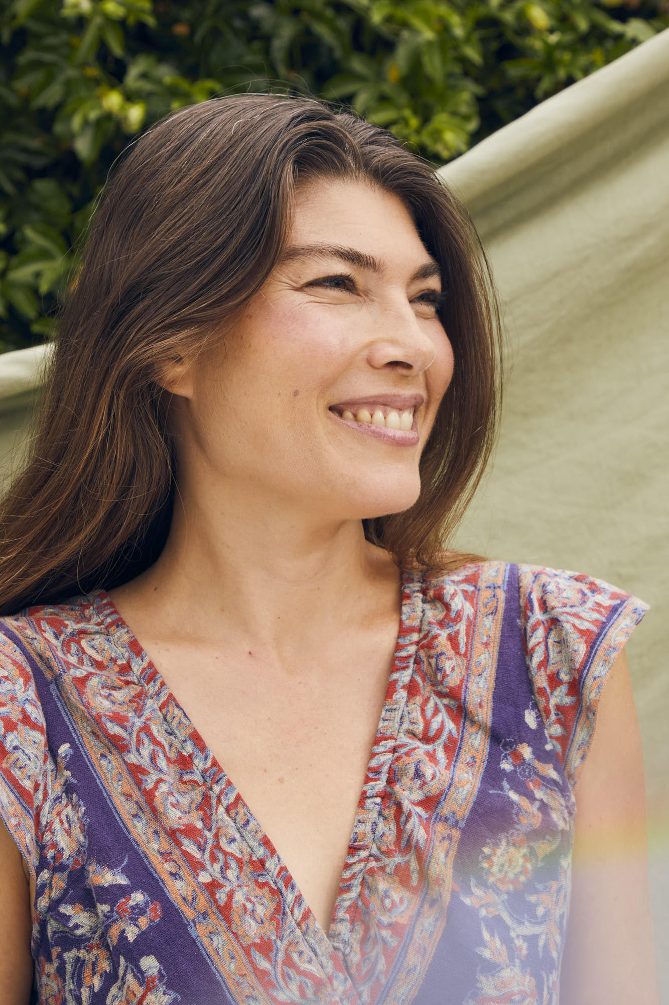 Smiling woman with long brown hair outdoors, wearing a colorful patterned top, in front of green foliage and a light-colored fabric