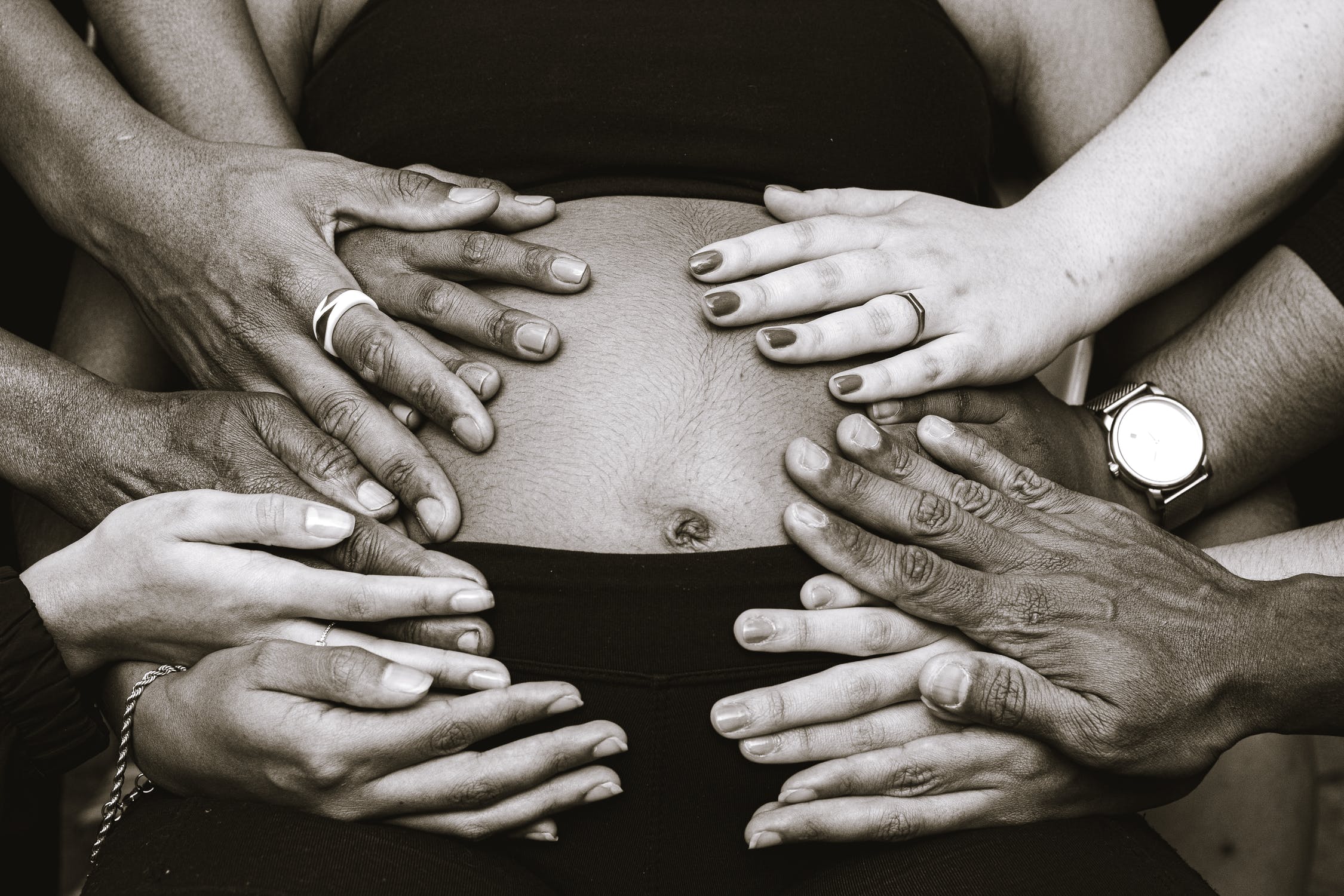 Multiple hands placed on a person's abdomen, some wearing rings and a watch, in black and white.