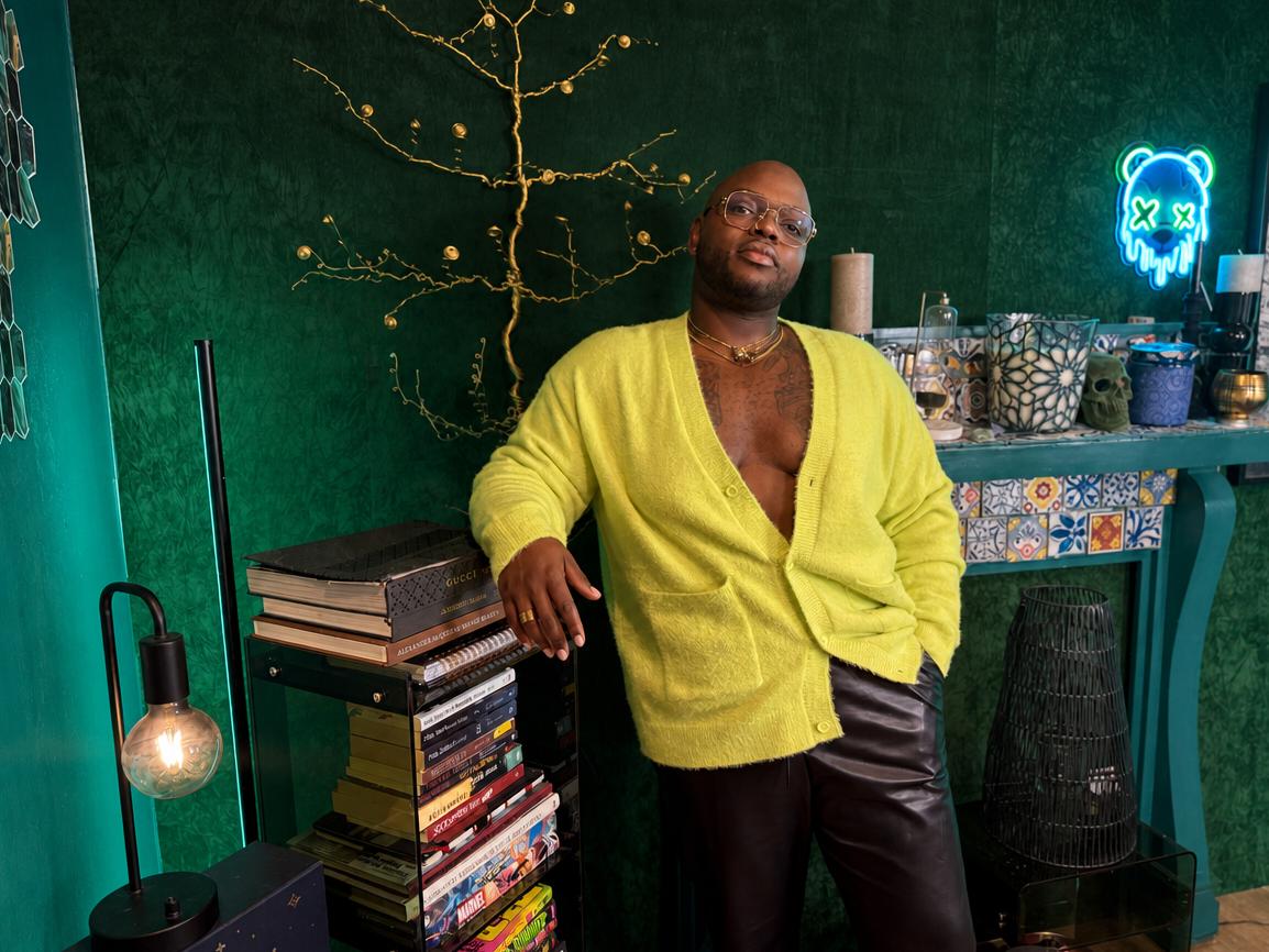 Person with glasses and a yellow cardigan standing in a room with green walls, books, and decorative items.