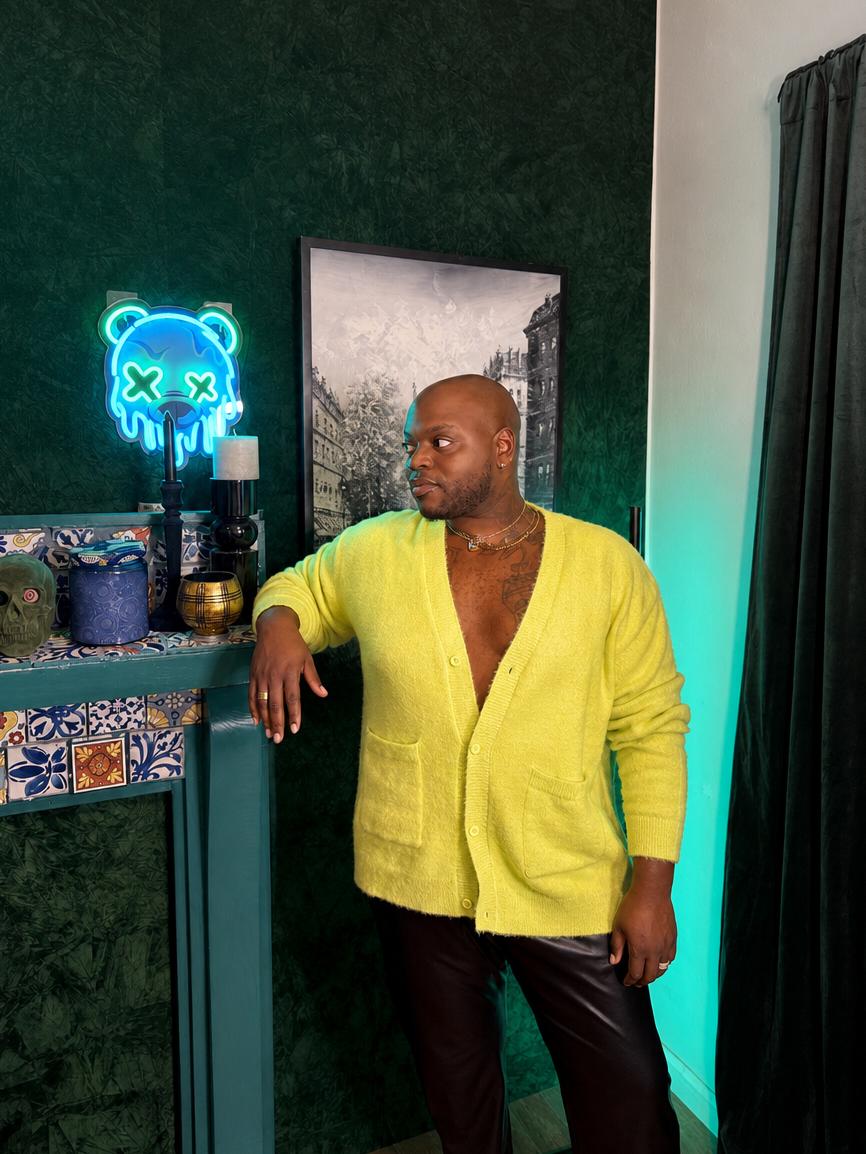 Man in yellow cardigan standing indoors near a decorated table and a neon bear sign, with a framed picture behind him.
