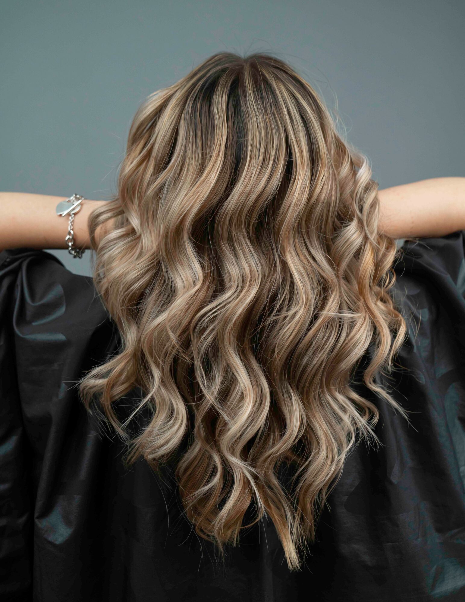 Back view of a woman with long, wavy blonde hair, wearing a black top and a bracelet, against a plain background.