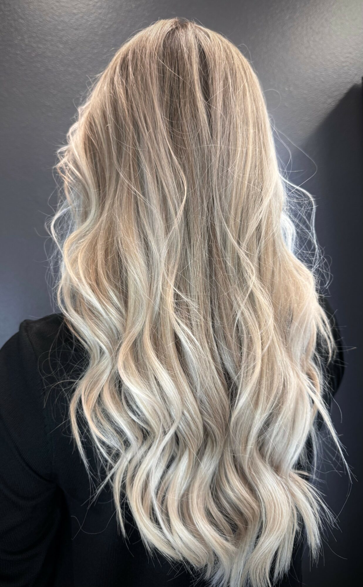 Back view of long, wavy blonde hair with darker roots, against a dark background.