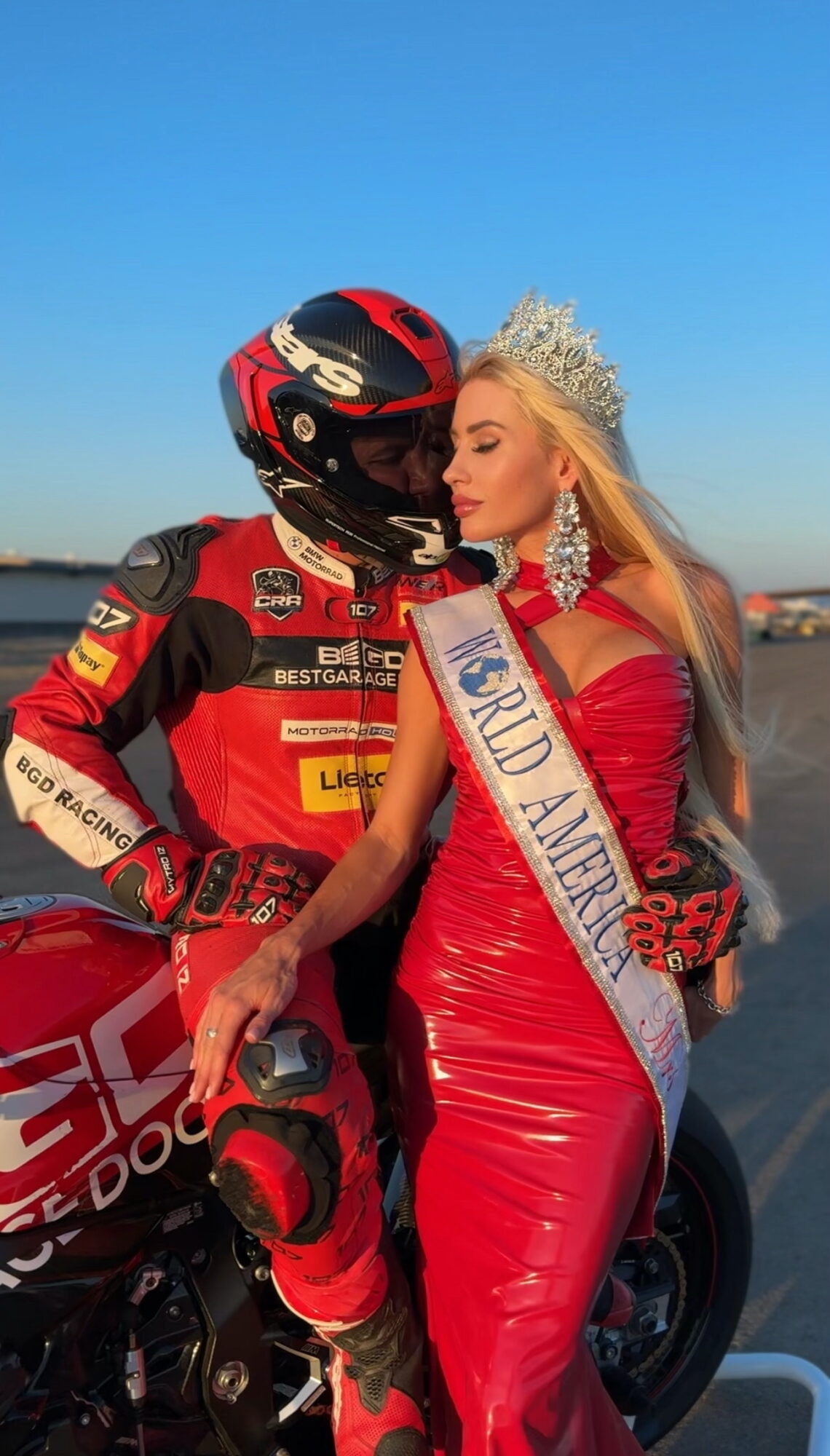 A woman in a red dress and sash stands close to a person in a racing suit and helmet, with a motorcycle nearby.