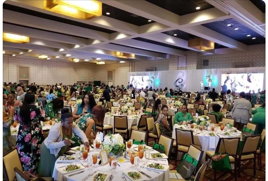 Large banquet hall filled with round tables, people, and a stage with screens, decorated for an event.