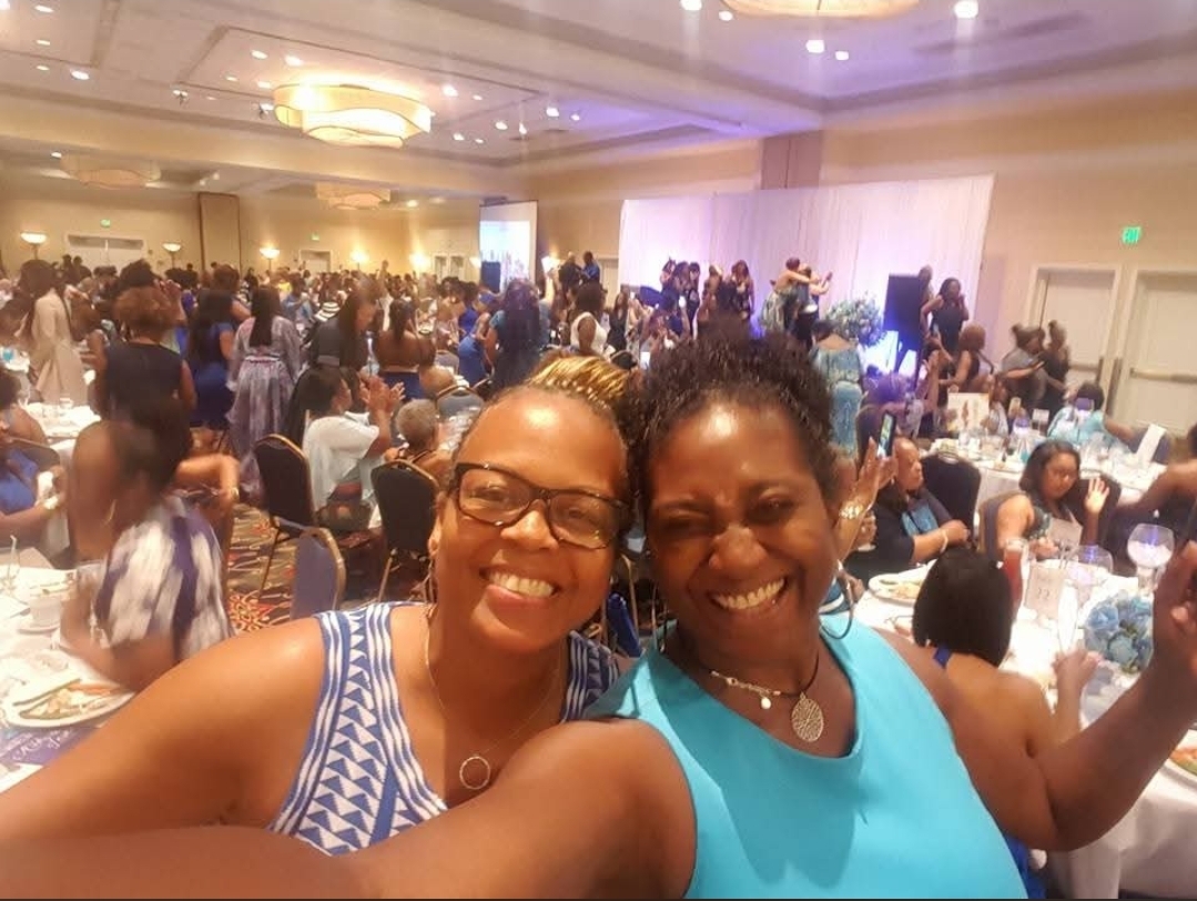 Two women smiling and taking a selfie at a large indoor event with many people and tables in the background.