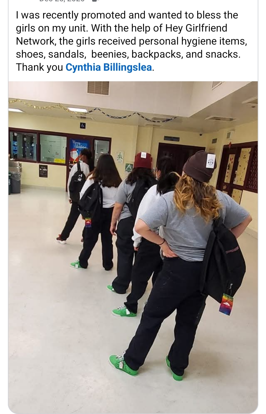 Group of women standing in line in a hallway, wearing casual clothes and green shoes, with a bulletin board and screens in the background.