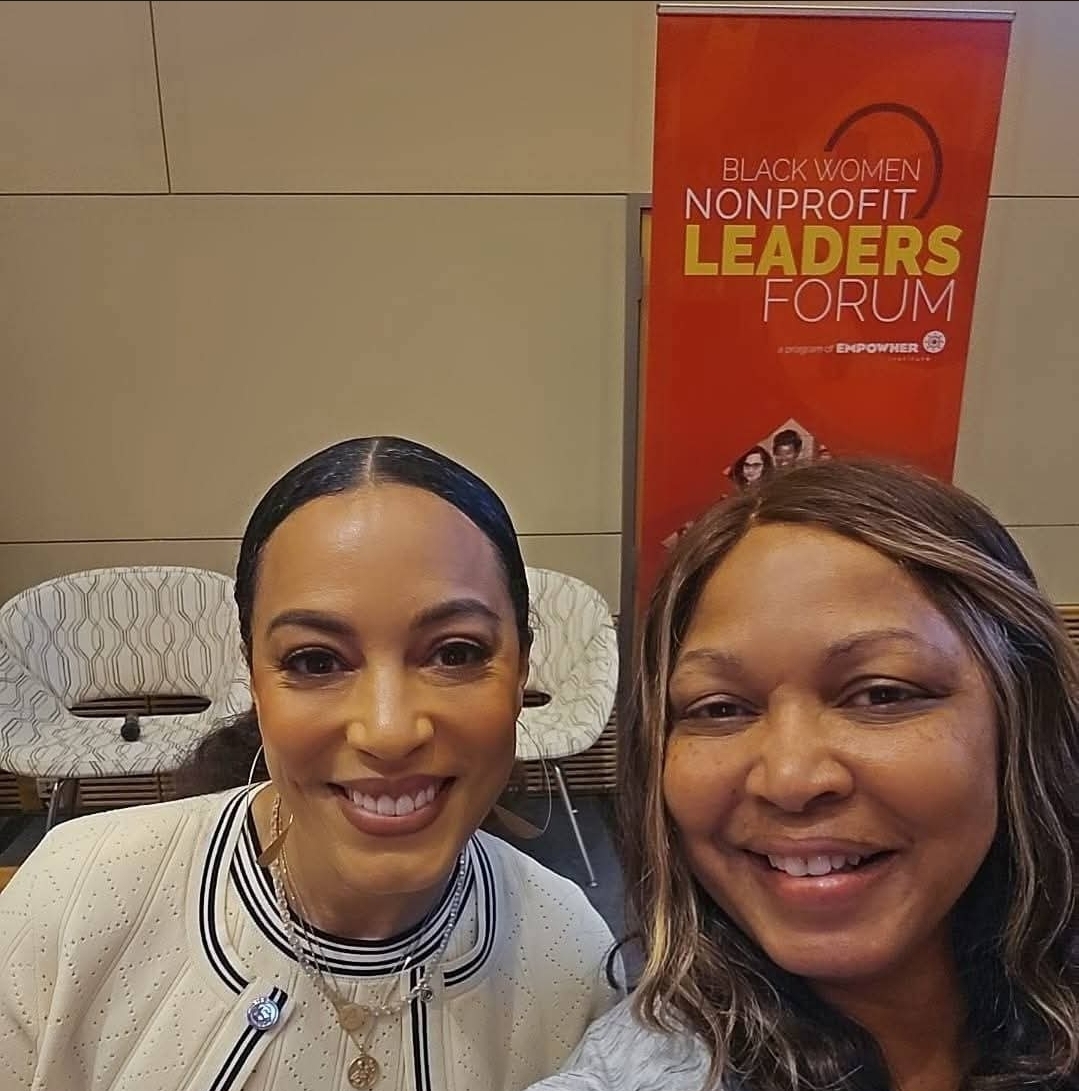 Two women smiling in front of a red banner that reads 'Black Women Nonprofit Leaders Forum'.