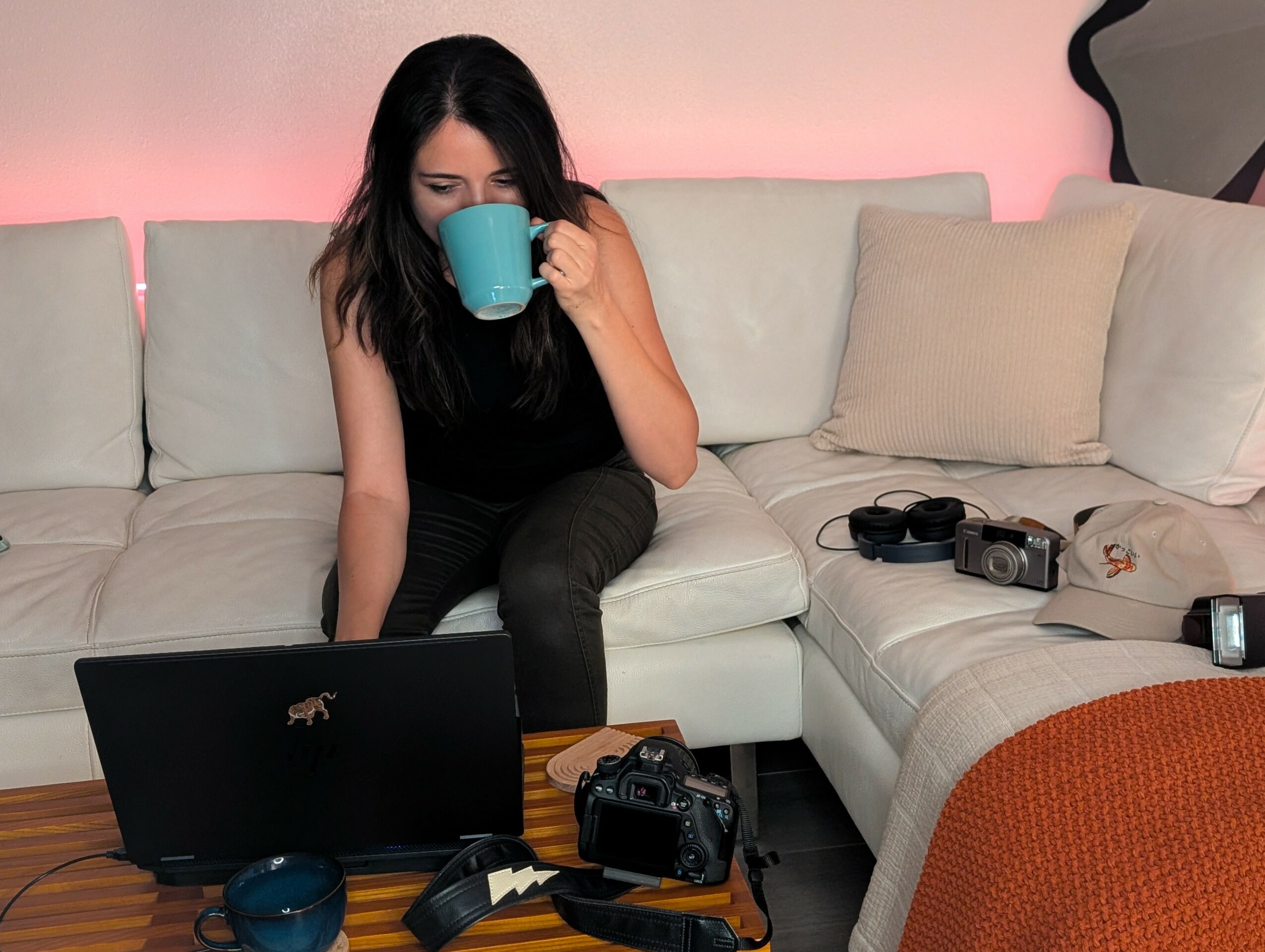 Woman sitting on a sofa drinking from a mug, with a laptop, camera, and headphones on the table in front.