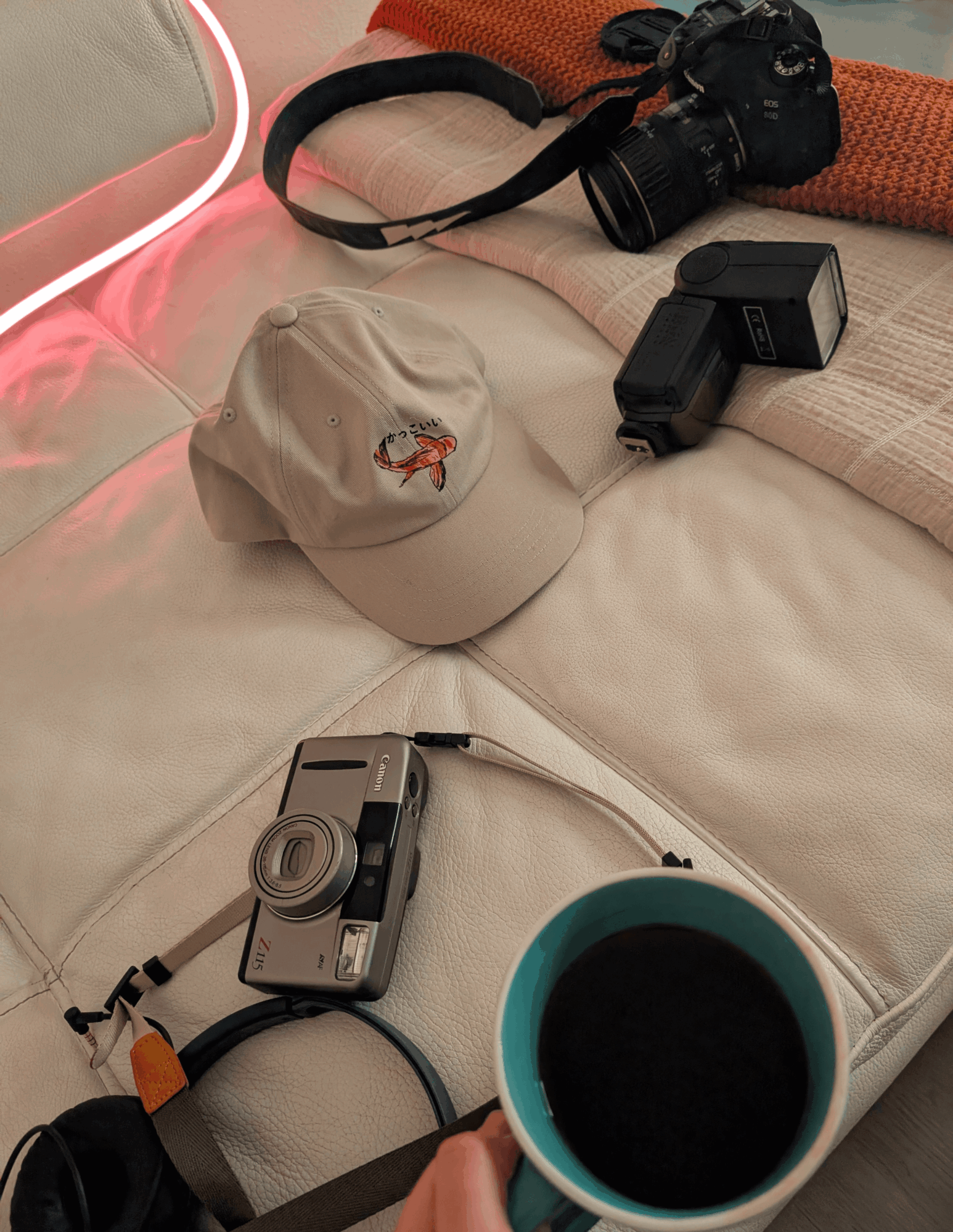 Camera, hat, and coffee cup on bed with camera gear in background.