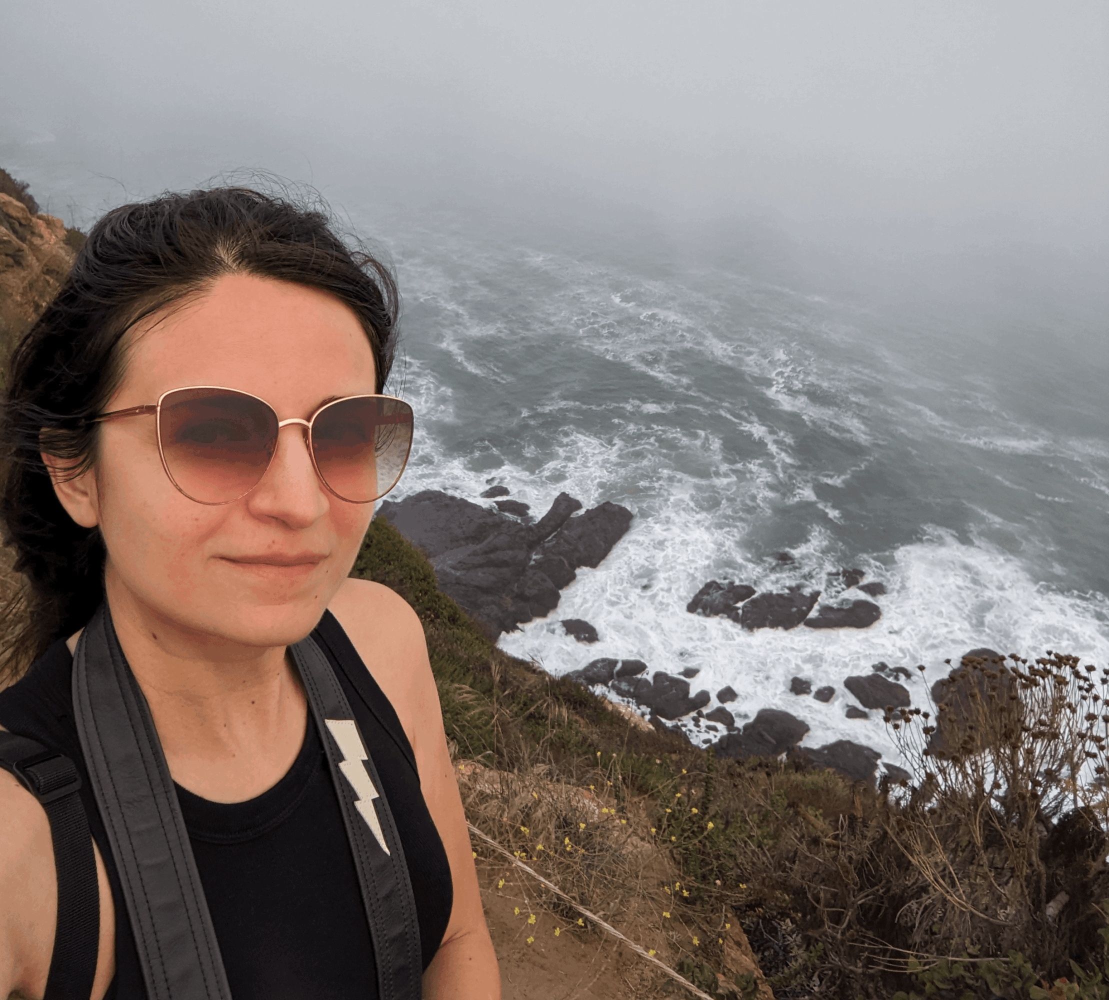 Woman with sunglasses and backpack near rocky coastline with misty ocean background.