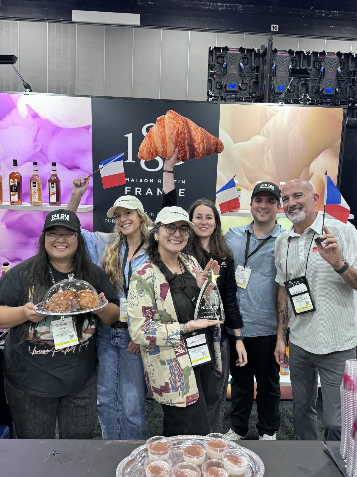 Group of five people smiling, holding food and drinks, standing in front of a colorful display at an event, with a large croissant prop above them.