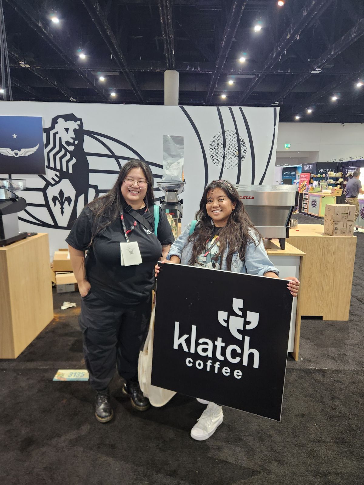 Two women standing indoors, holding a large black sign with white text that reads 'klatch coffee'.