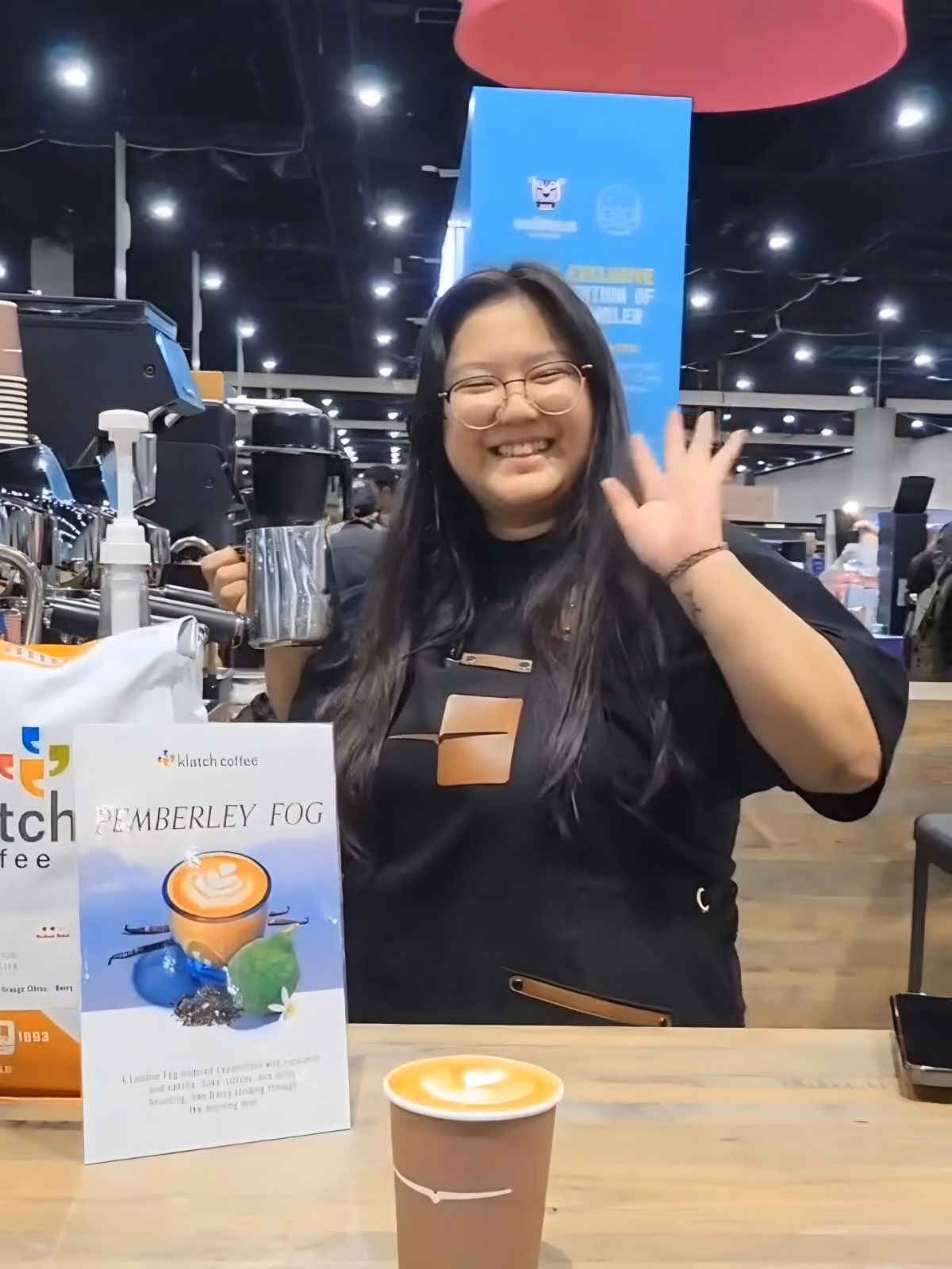 Smiling woman with glasses waving at camera in a store, surrounded by coffee products and display signs.