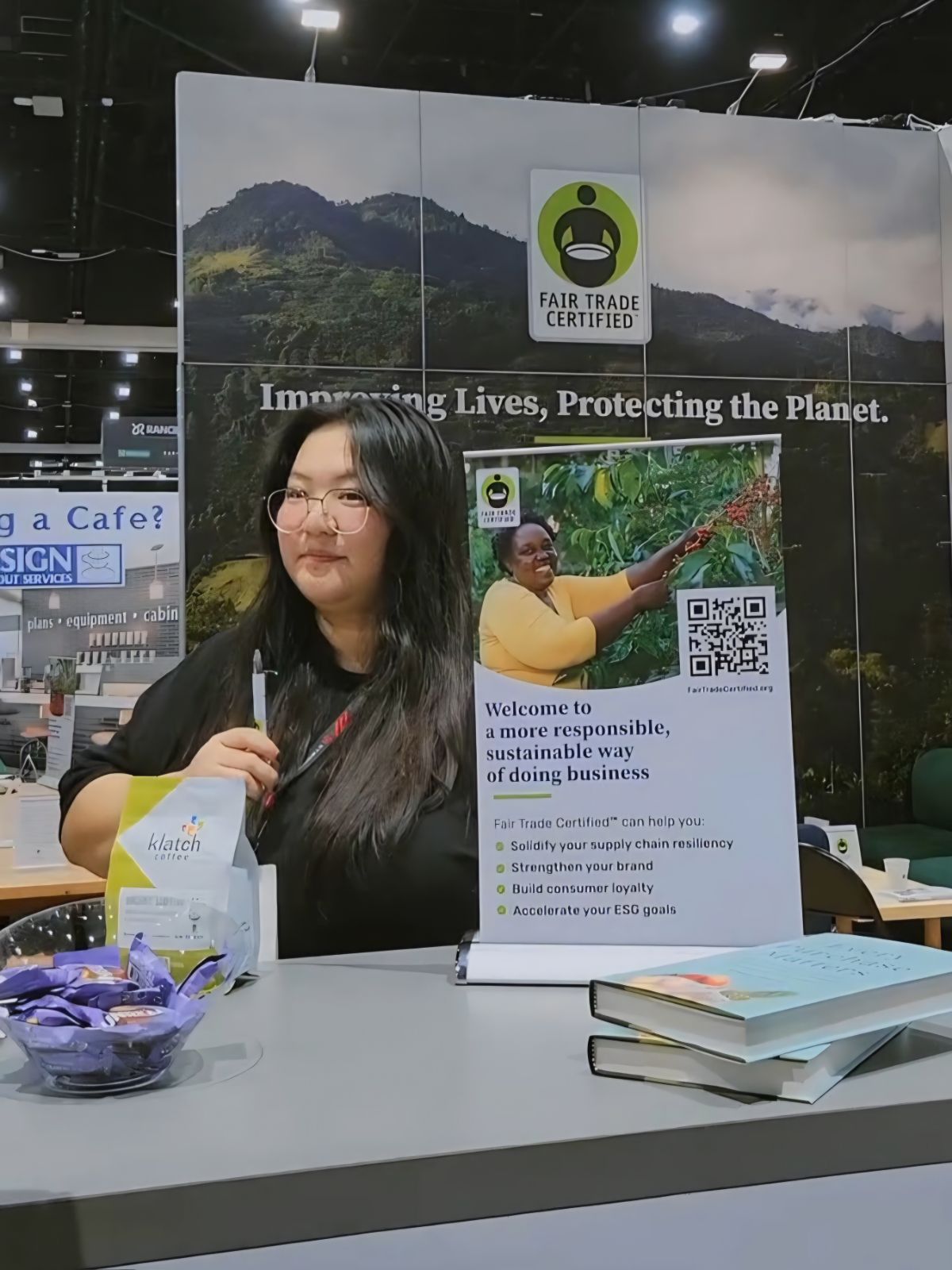 Woman with long dark hair and glasses standing behind a table with brochures and a bowl, in front of a banner with mountains and a logo.