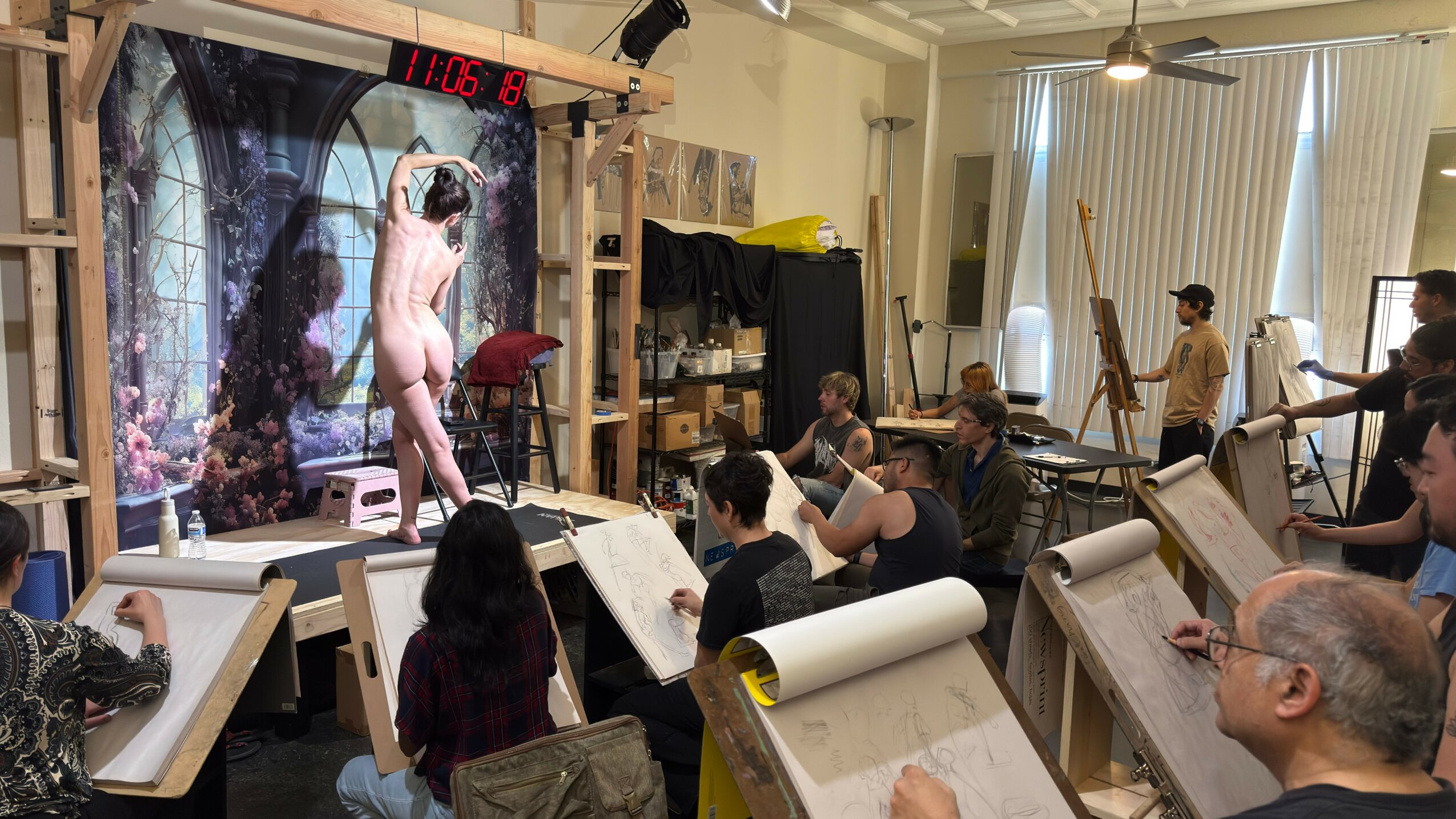 People in a classroom watching a live nude model pose on a stage with a forest backdrop, some taking notes.