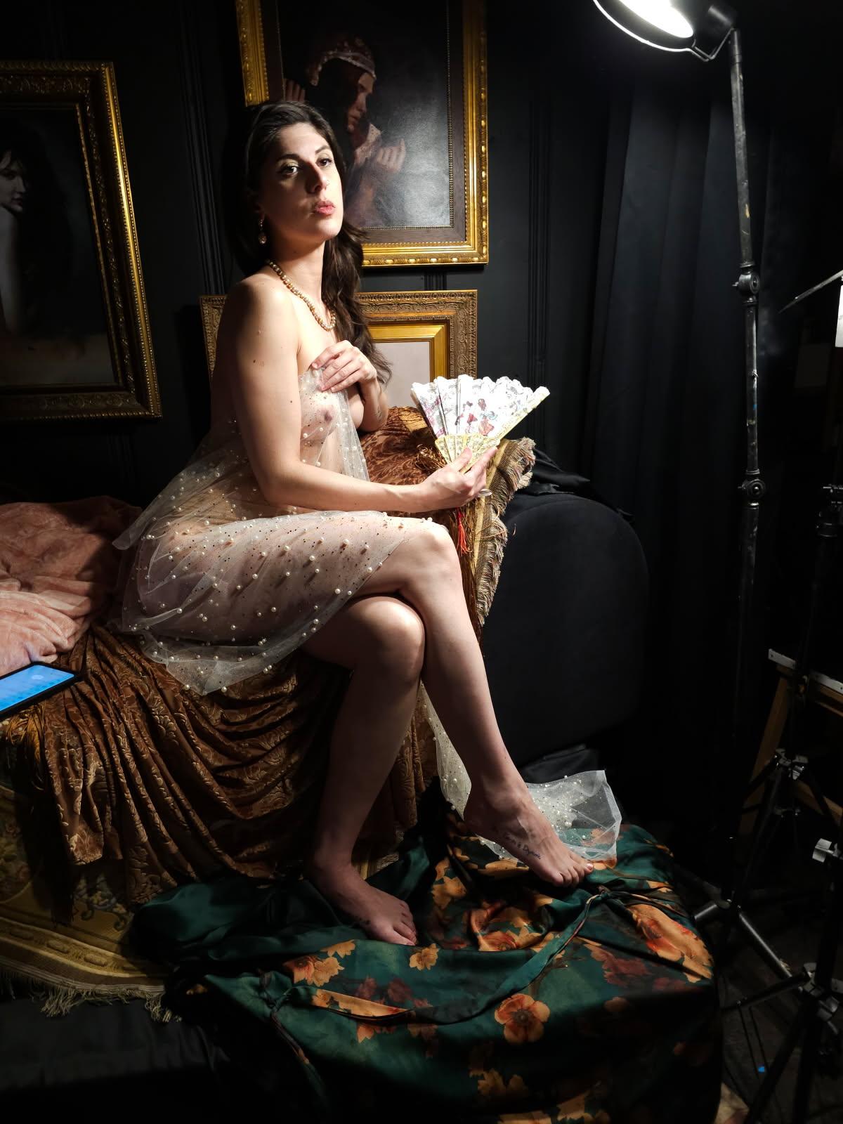 Woman sitting on a bed holding a fan, in a dimly lit room with framed paintings, wearing a light dress.